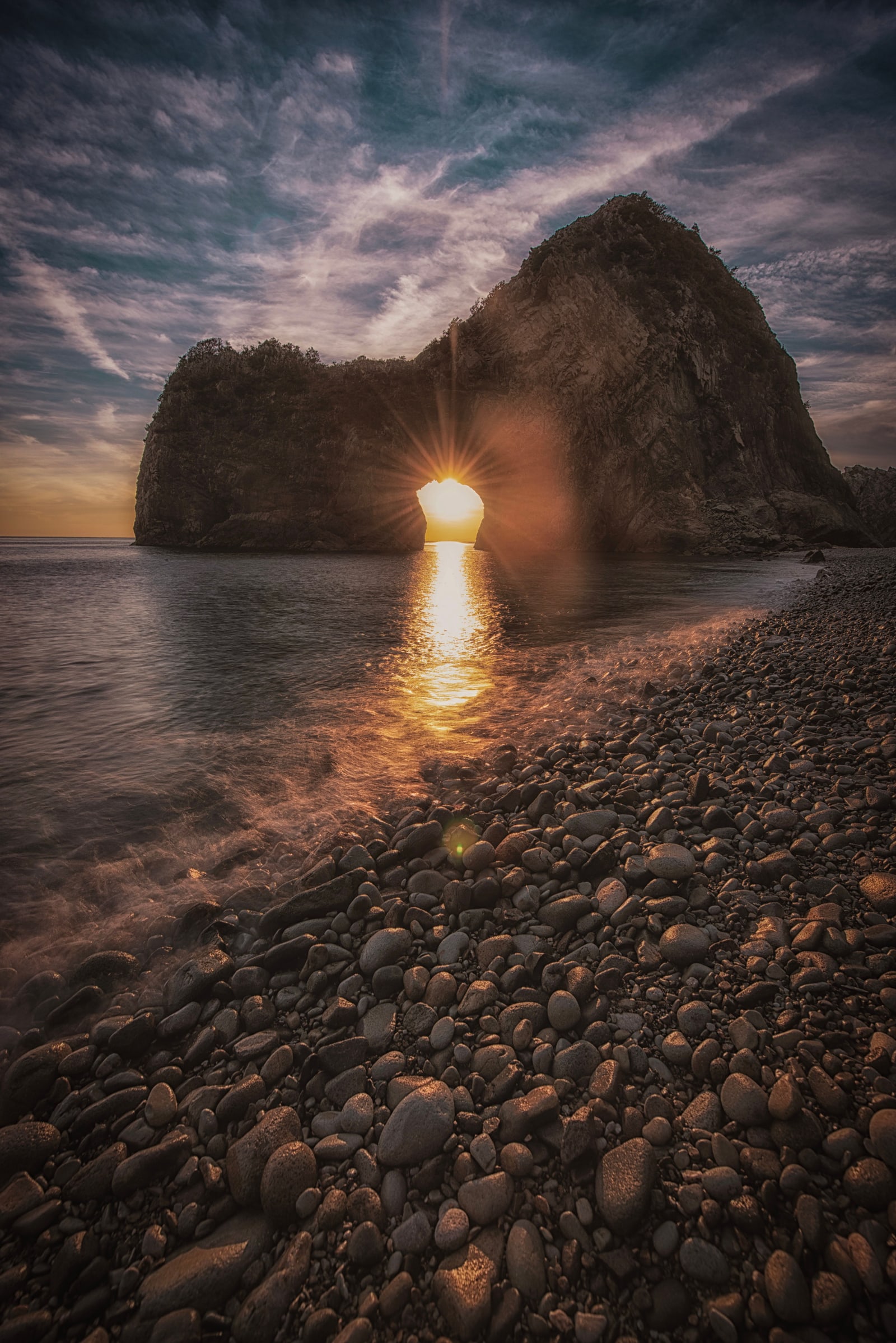 The sun setting through a rock arch, seen from a pebble beach in Izu, as the photographer places it