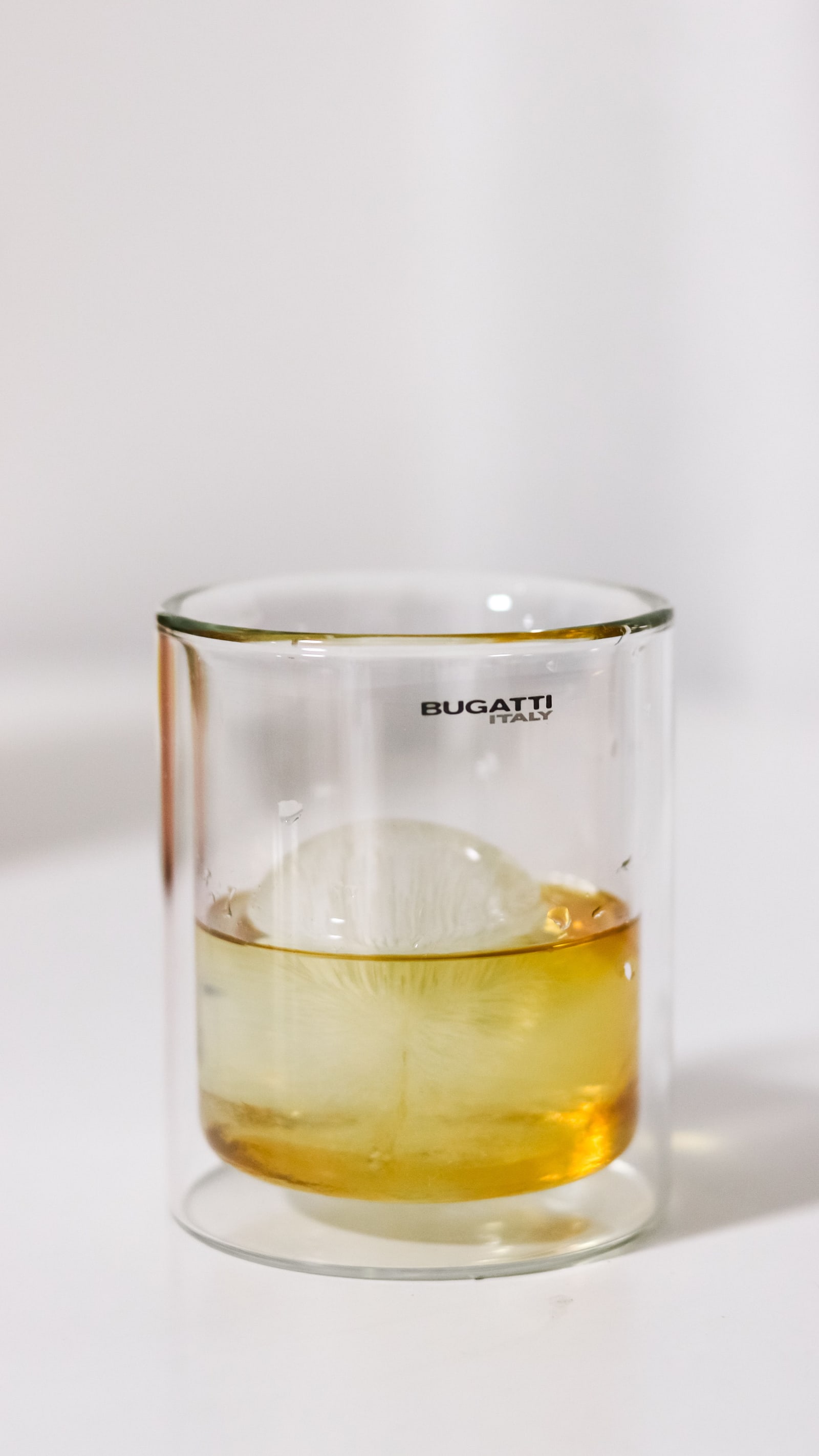 An ice sphere in a double-walled glass of pale amber spirit on a white surface.