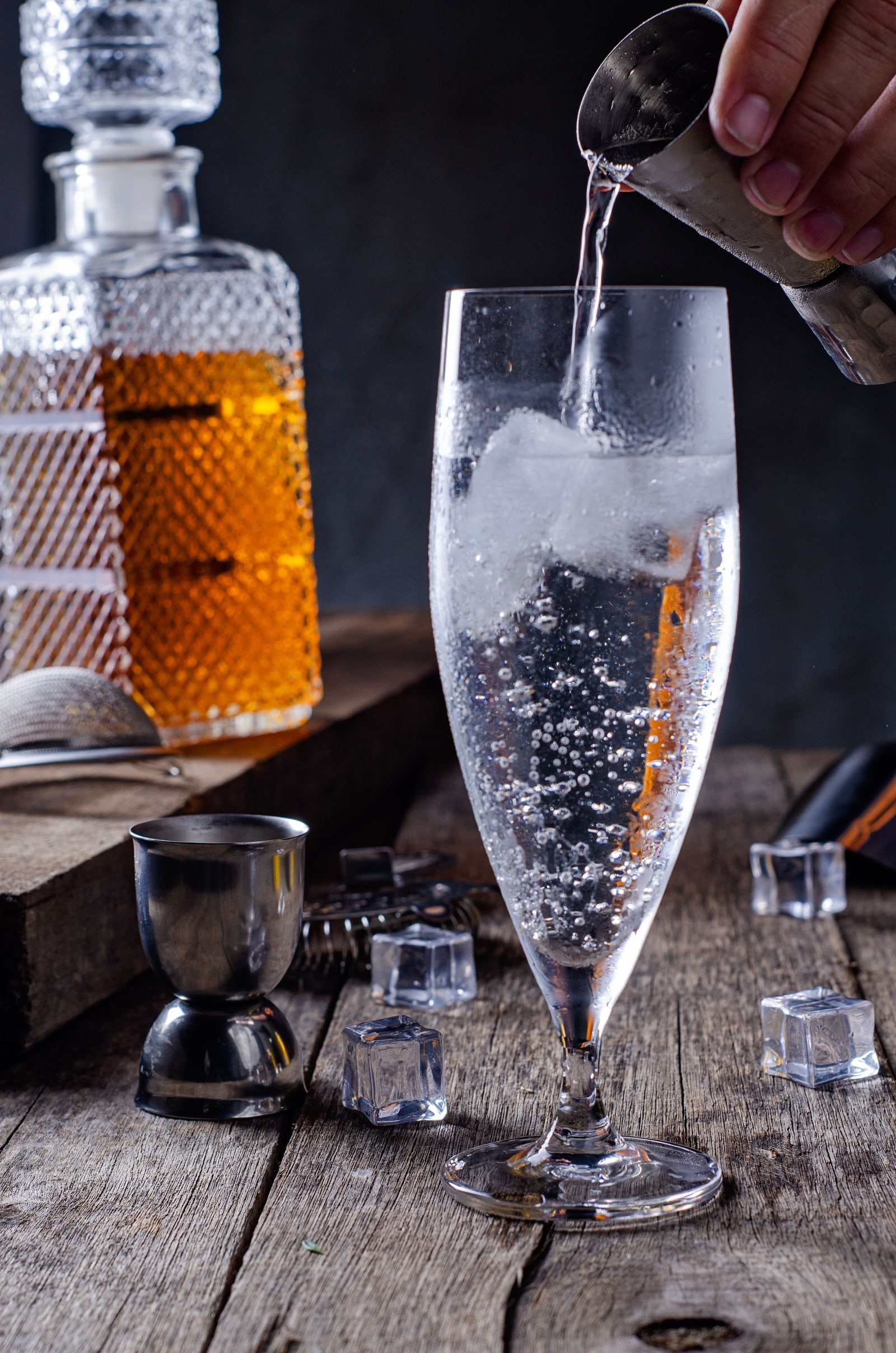 Soda being poured over ice into a tall glass, with a decanter of amber spirit out of focus behind.