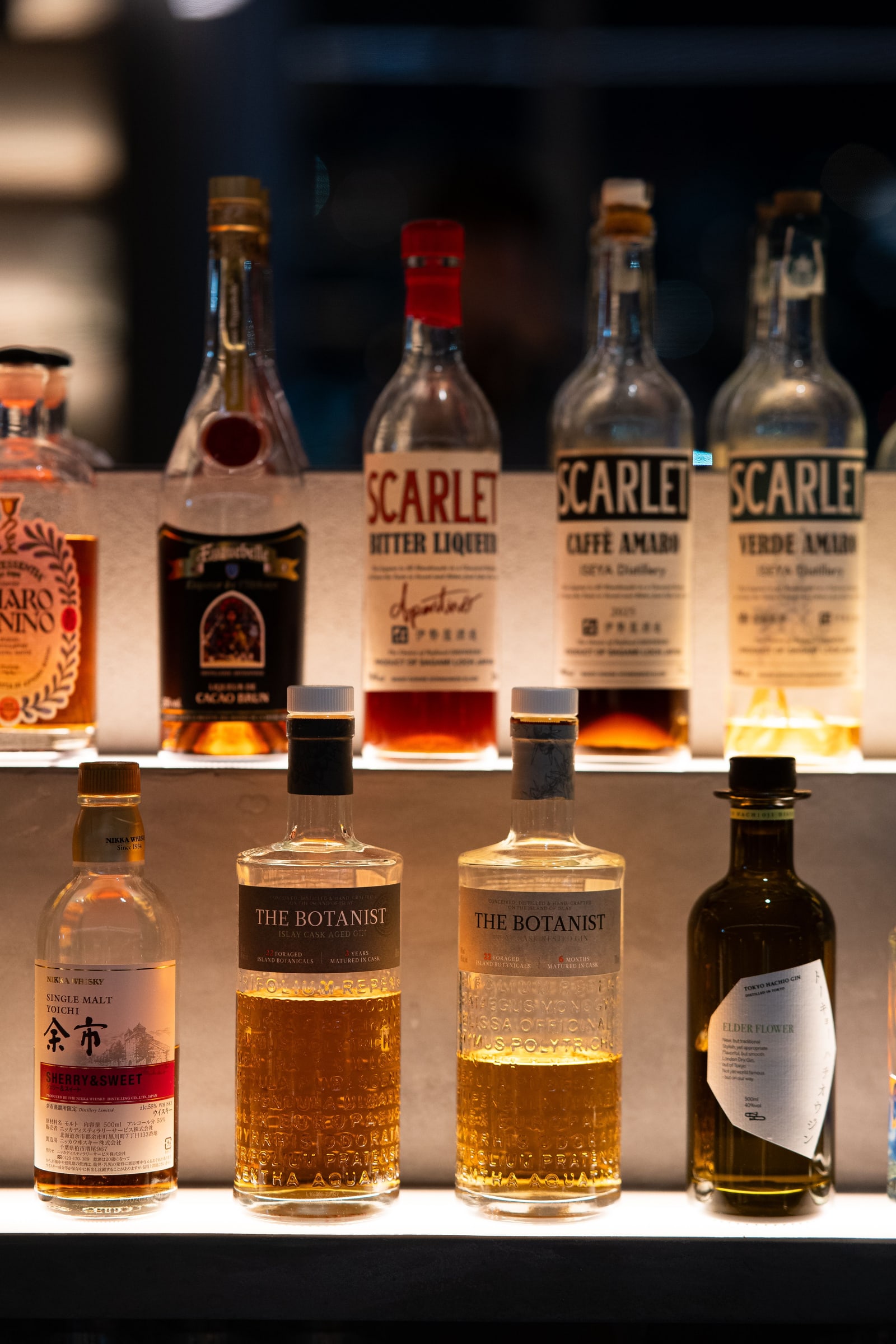 A lit two-tier bar shelf with liqueurs, two gins and one Nikka Yoichi single malt.