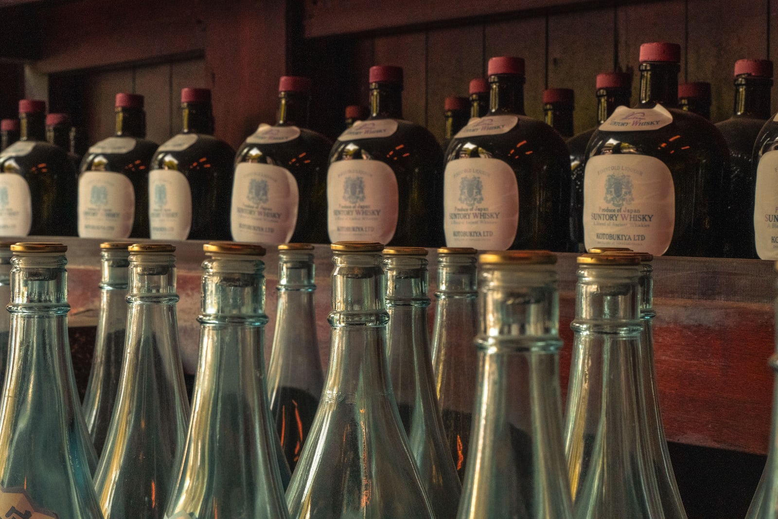 Rows of vintage Suntory whisky bottles with Kotobukiya-era labels on a wooden bar shelf, empty clear bottles in front.