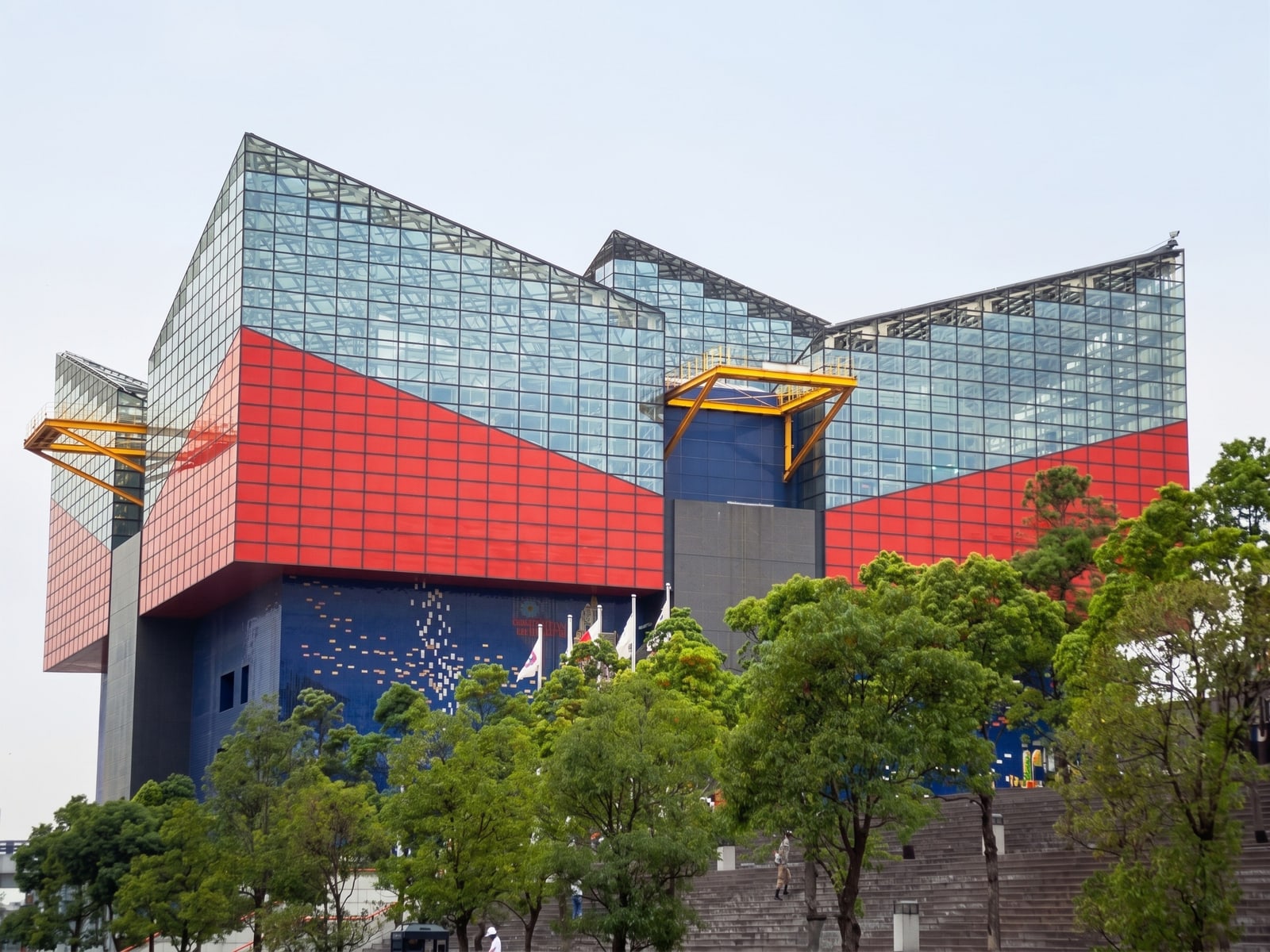 The Kaiyukan aquarium building, red and blue panels under glass peaks, with trees and steps in front.