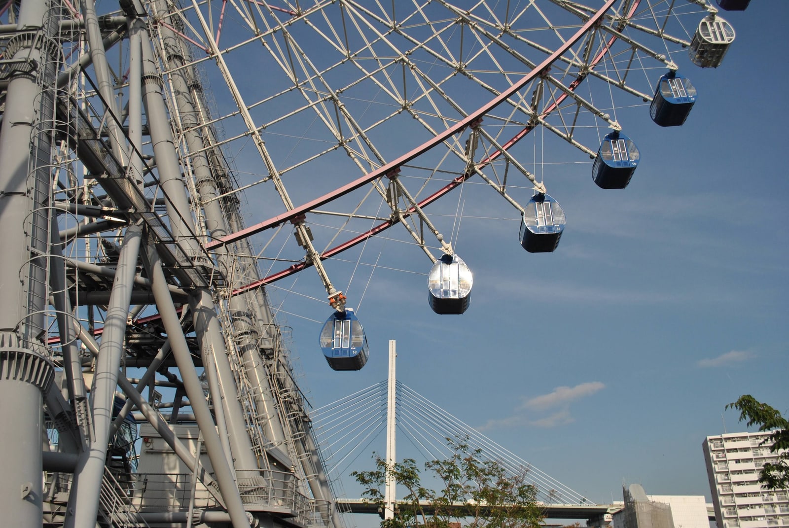 Gondolas on the steel frame of a giant Ferris wheel in Osaka, a bridge tower behind it under blue sky.