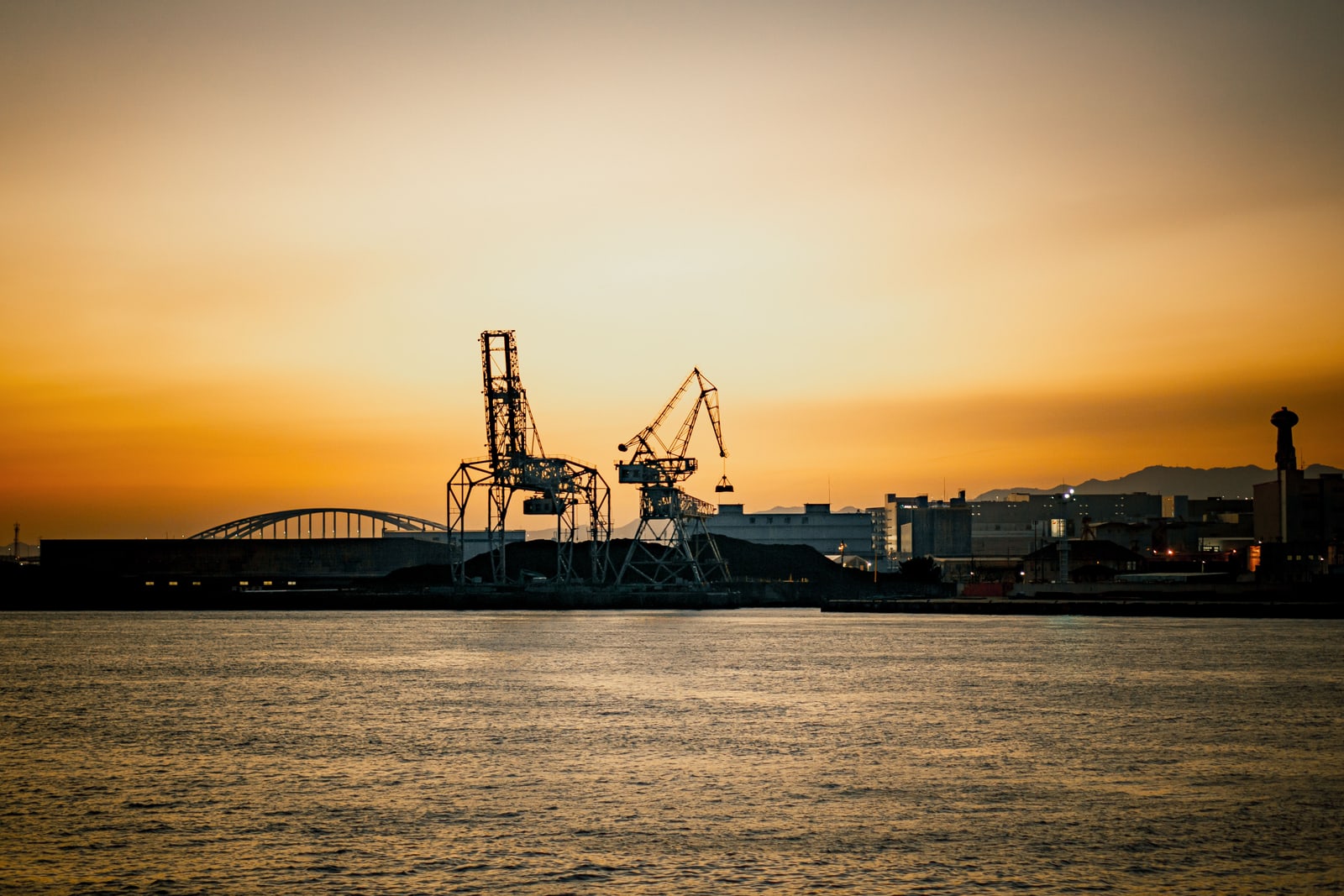 Port cranes and a bridge silhouetted against an orange sunset over the water of Osaka Harbor.