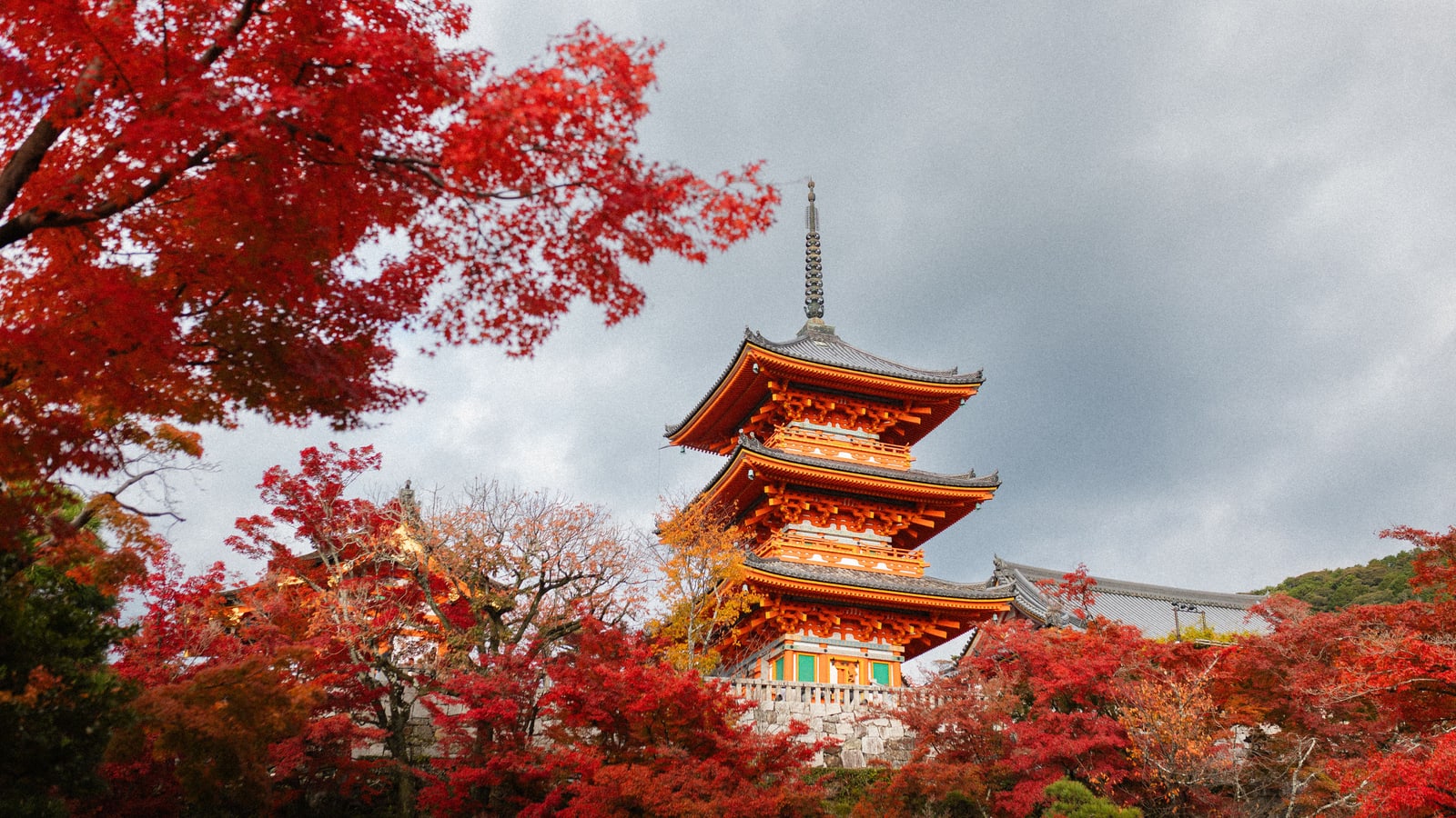 A vermilion three-story pagoda under a grey sky, framed by red maple branches, with red trees below it.