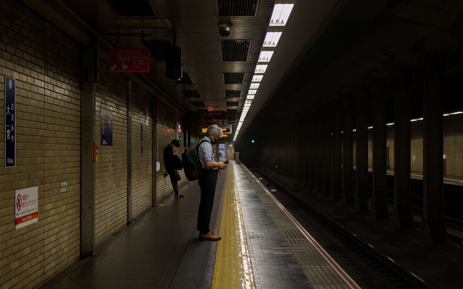 A dim, tiled Kyoto subway platform with a yellow tactile strip, two people waiting for a train.