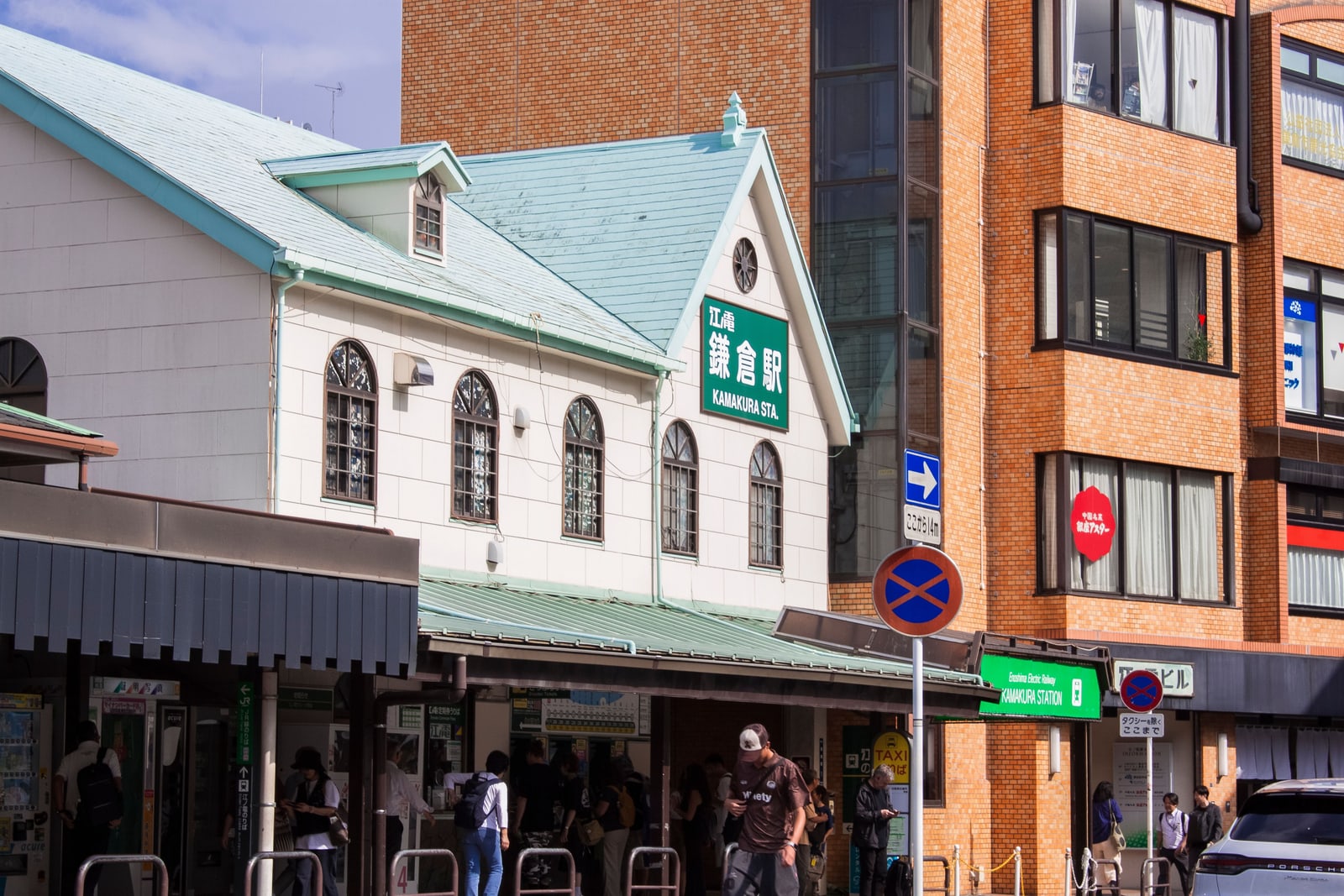 The front of Enoden Kamakura Station, a white building with a pale green roof and a green sign reading Kamakura Station, commuters at the ticket machines below.