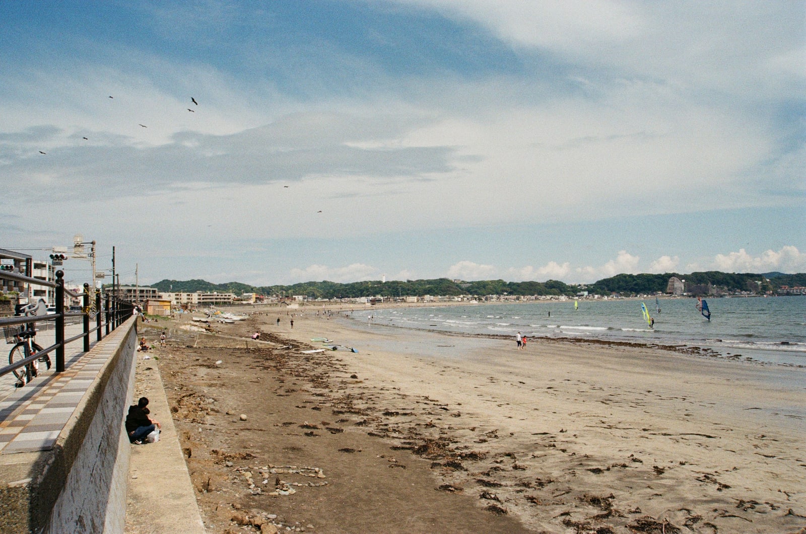 A long sand beach curving along a bay under broken cloud, windsurf sails on the water and a few walkers on the sand.