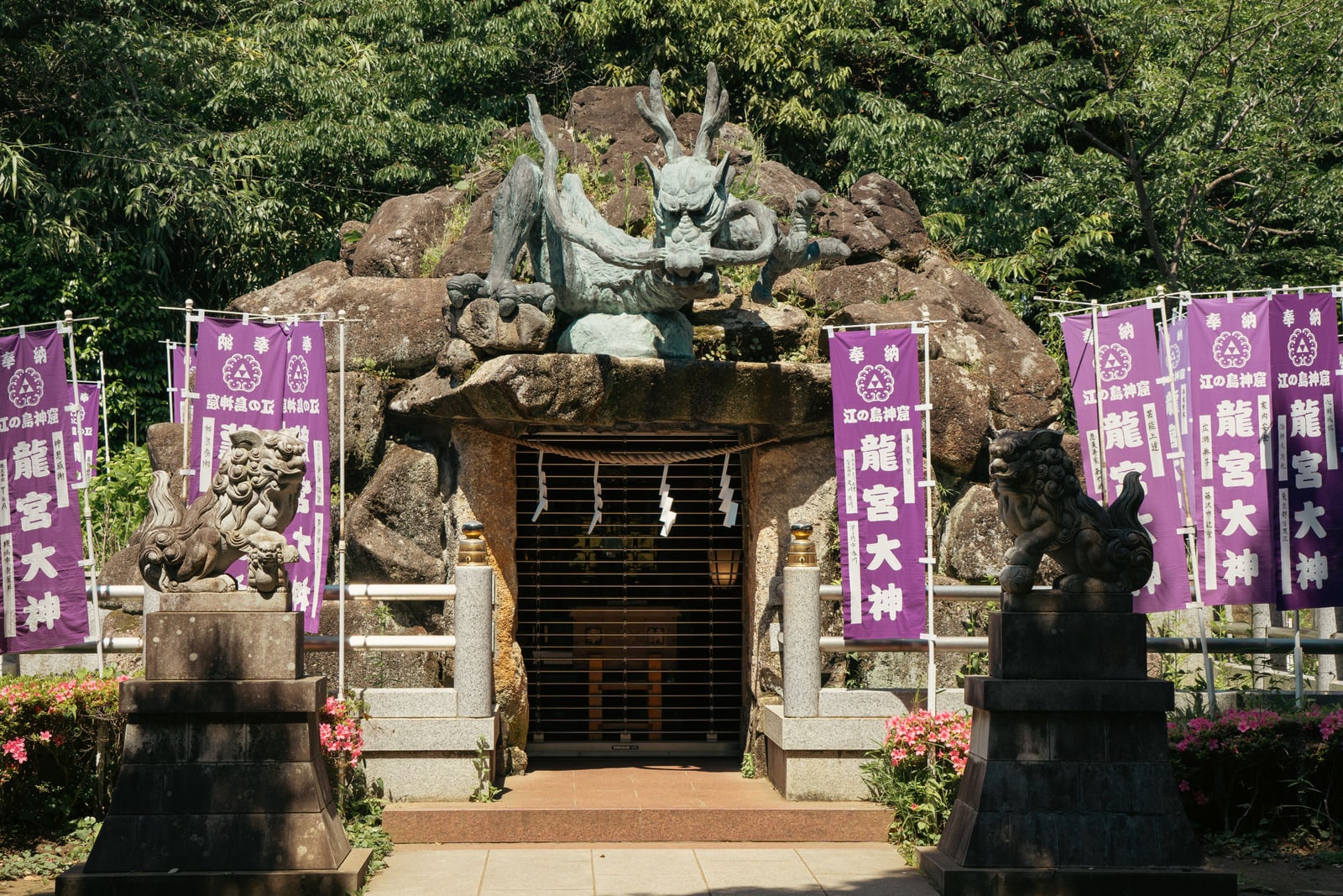 A bronze dragon above the cave shrine entrance on Enoshima, stone guardian lions on either side and purple banners reading the shrine cave and Ryugu Daijin.