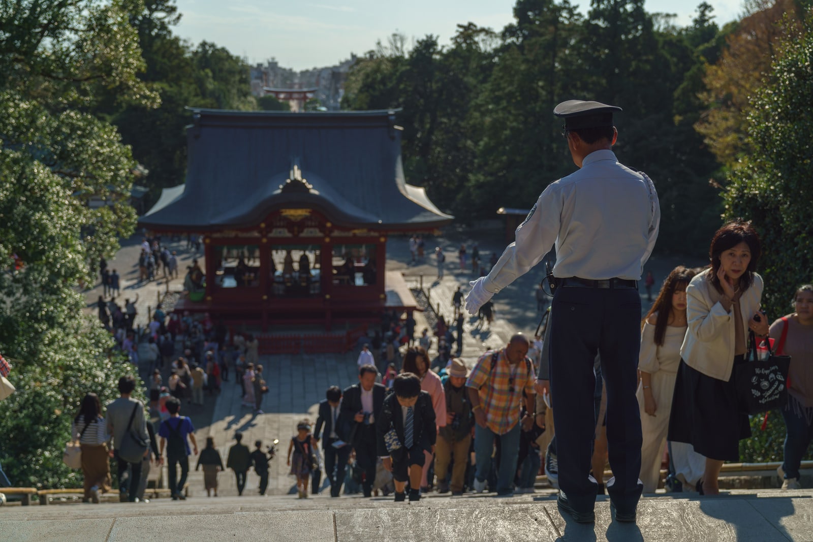 Visitors coming up a wide flight of stone steps at Tsurugaoka Hachimangu, a guard in uniform at the top, a red-framed hall with a grey tiled roof below and a red torii far down the approach.