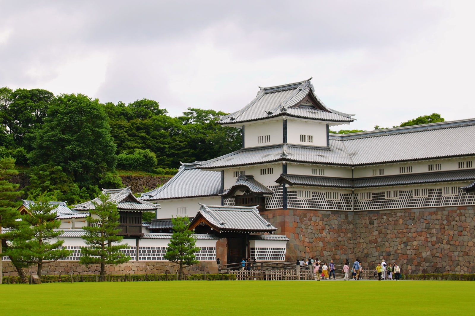 A white-walled gatehouse with grey tiled roofs above a stone wall at Kanazawa Castle Park, a wide lawn in front and visitors at the entrance
