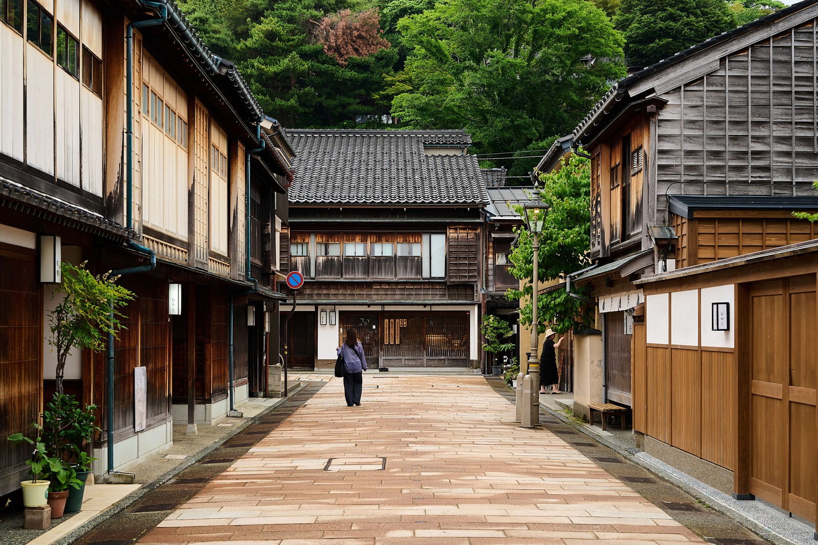 A quiet stone-paved lane between wooden townhouses with dark tiled roofs in Kanazawa, one woman walking away