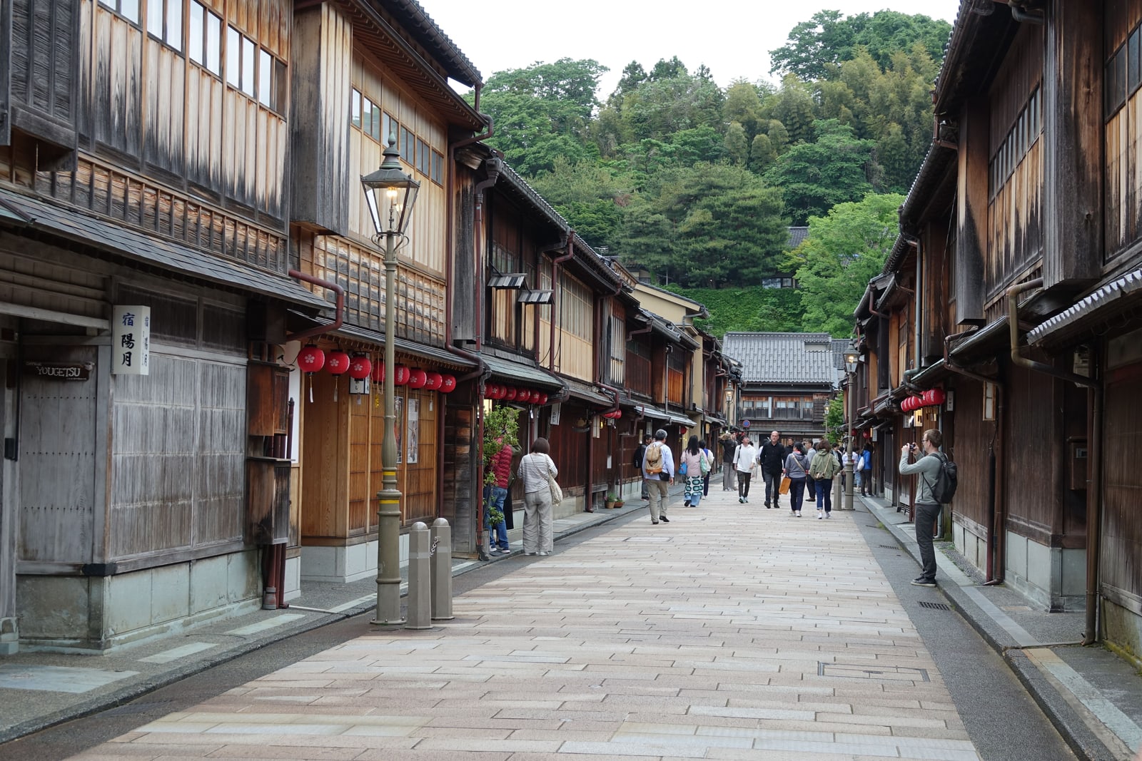 A wide stone-paved street between wooden townhouses with latticed fronts and red lanterns in the Higashi Chaya District, visitors walking