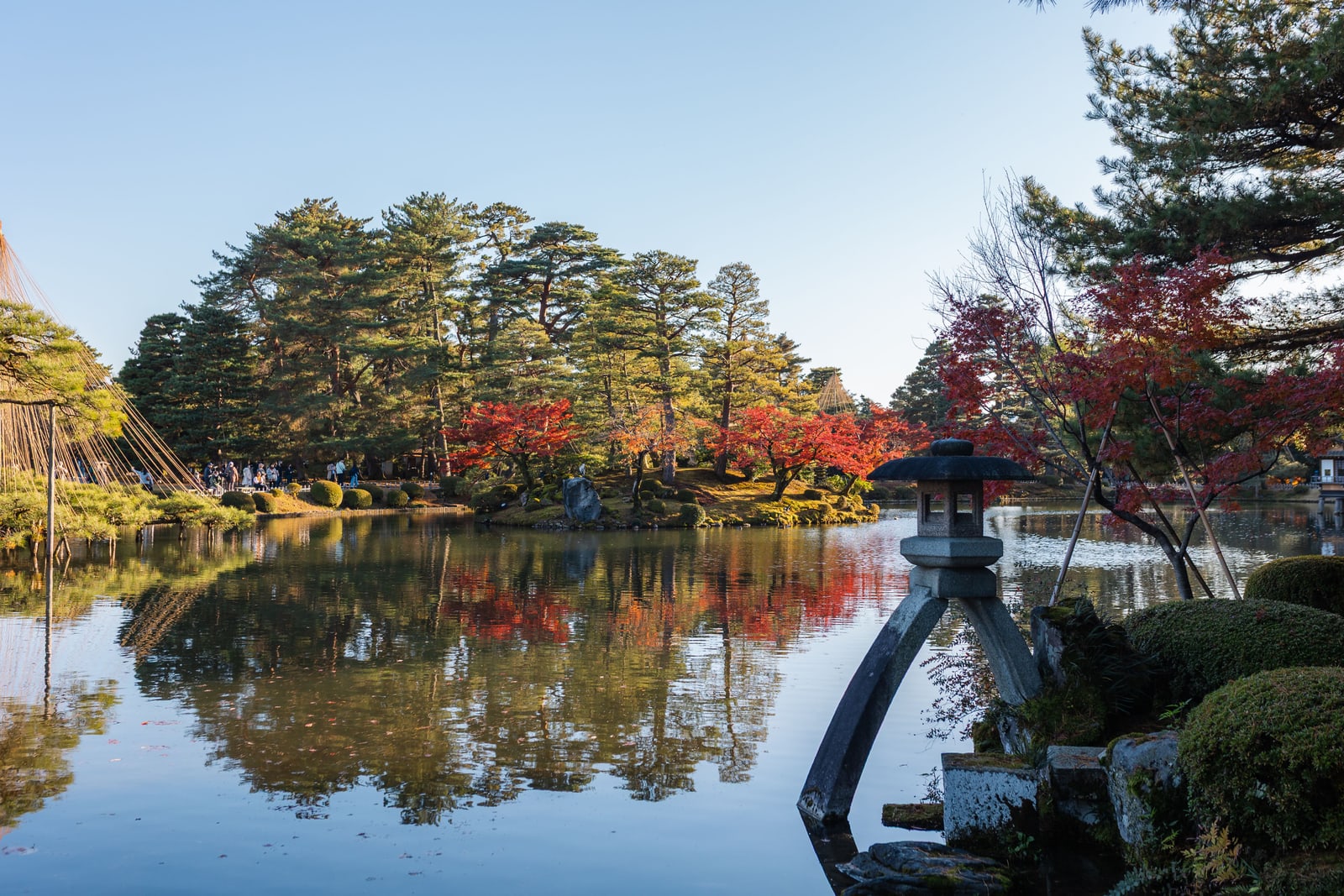 A still pond in Kenrokuen Garden reflecting pines and red maples, a stone lantern on two curved legs standing in the water at right