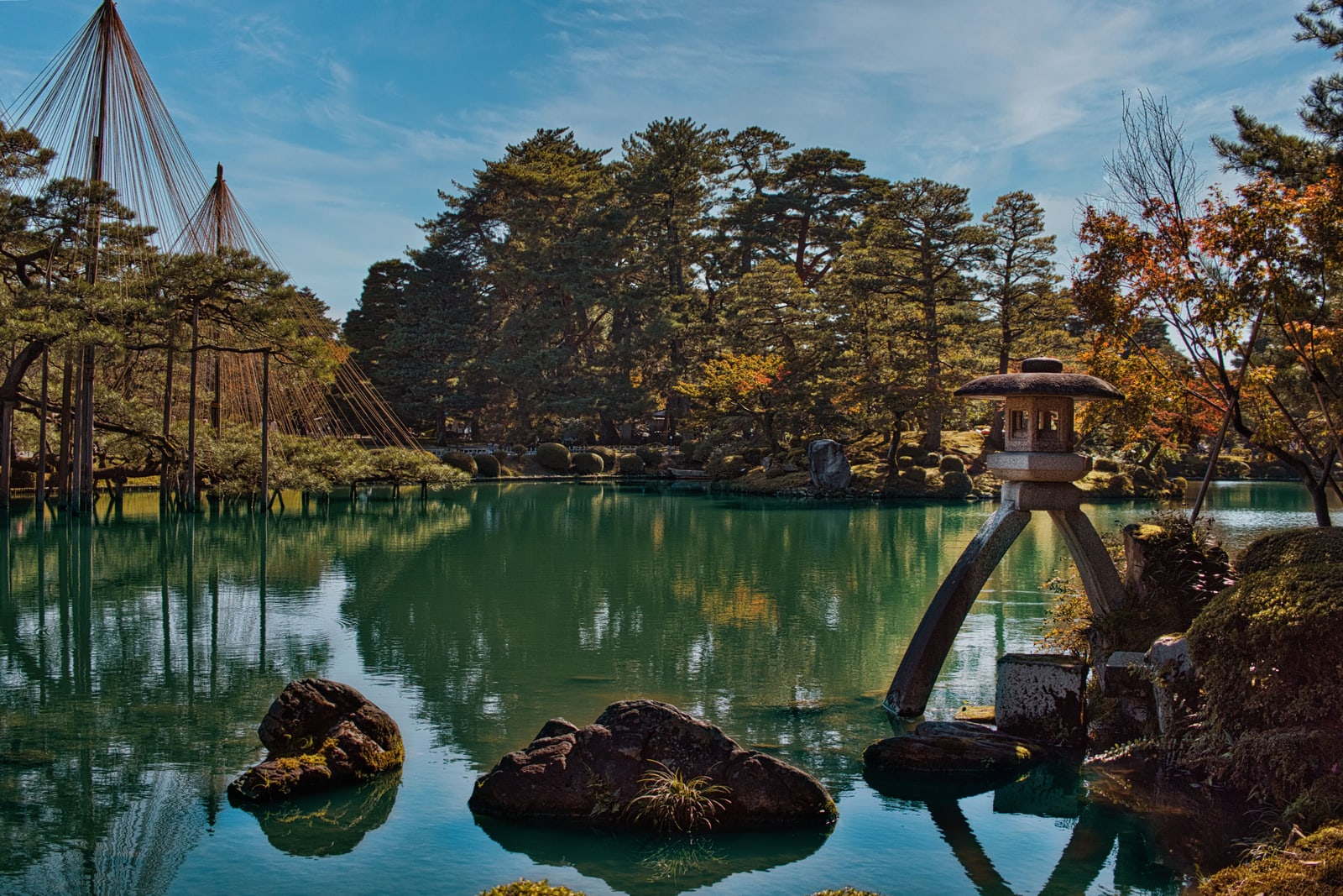 A two-legged stone lantern at the edge of a pond in Kenrokuen Garden, pines held up by rope supports on the left and autumn leaves on the right