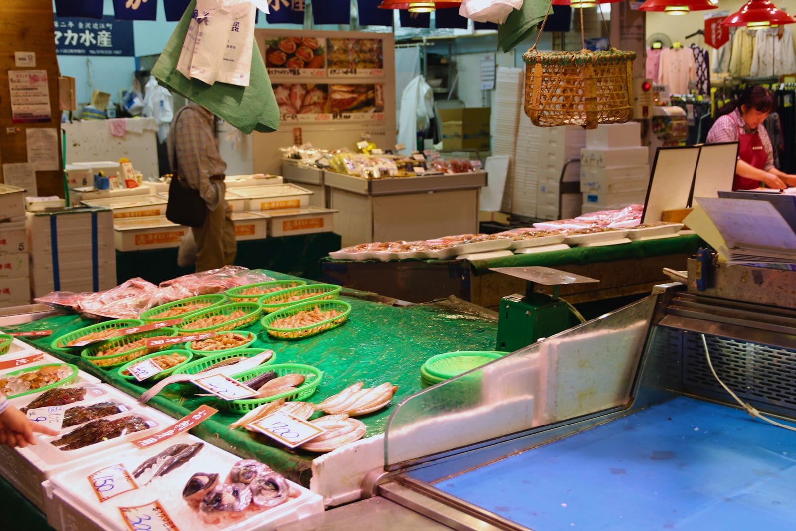 Trays of small shrimp and whole fish with hand-written price tags at a fish stall in Omicho Market, Kanazawa