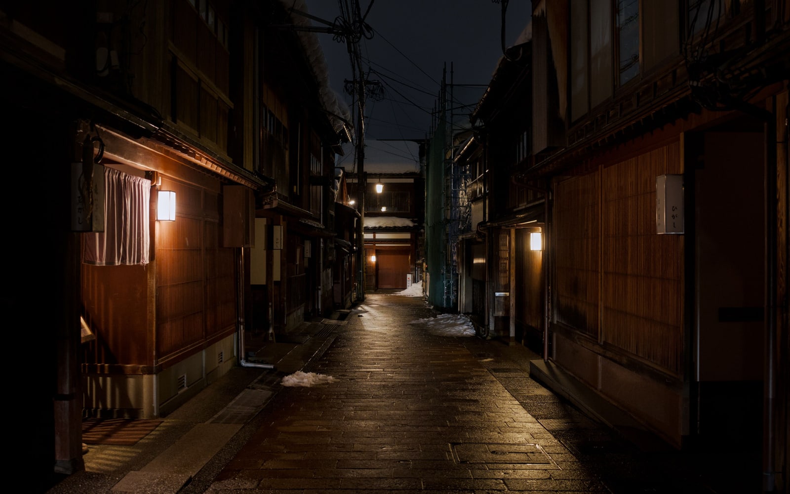 A narrow stone-paved alley of wooden buildings at night, wall lanterns glowing and patches of snow on the pavement