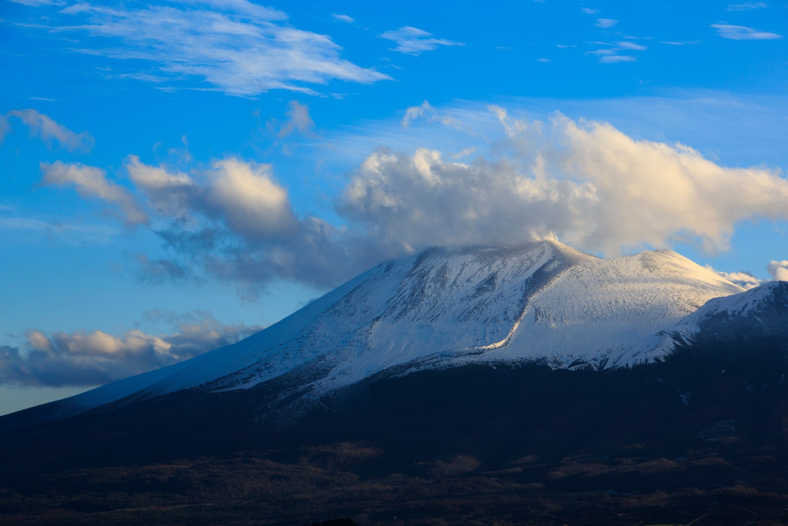 A broad snow-covered volcano with cloud over the summit under a blue sky