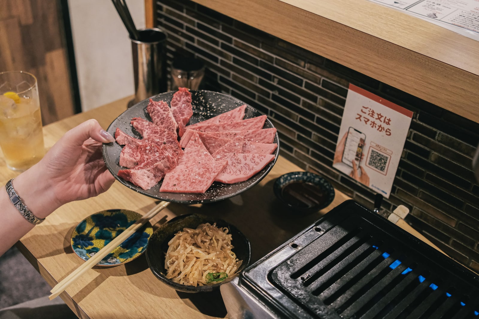 A hand holding a plate of marbled raw beef slices over a table grill, small side dishes and a drink beside it.