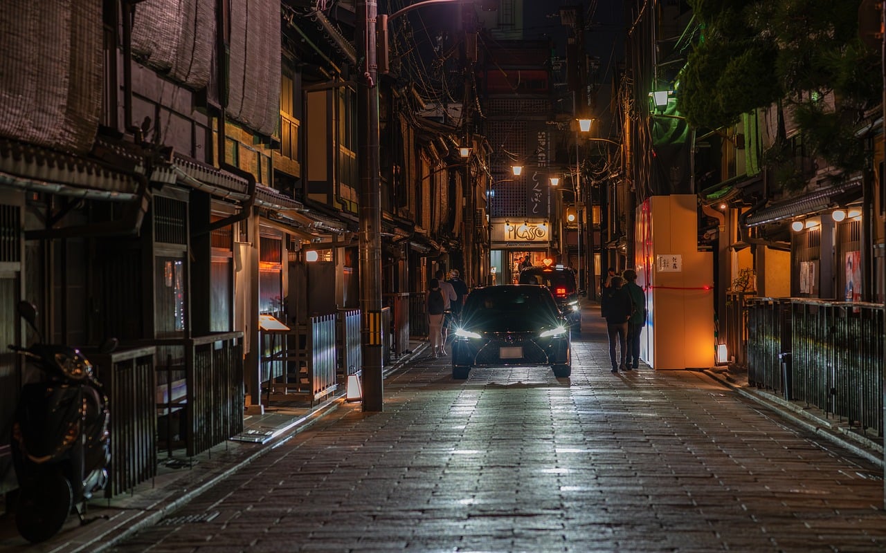 A stone-paved street at night between dark wooden townhouses with lattice fronts and lit lanterns, a car with headlights coming down the middle.