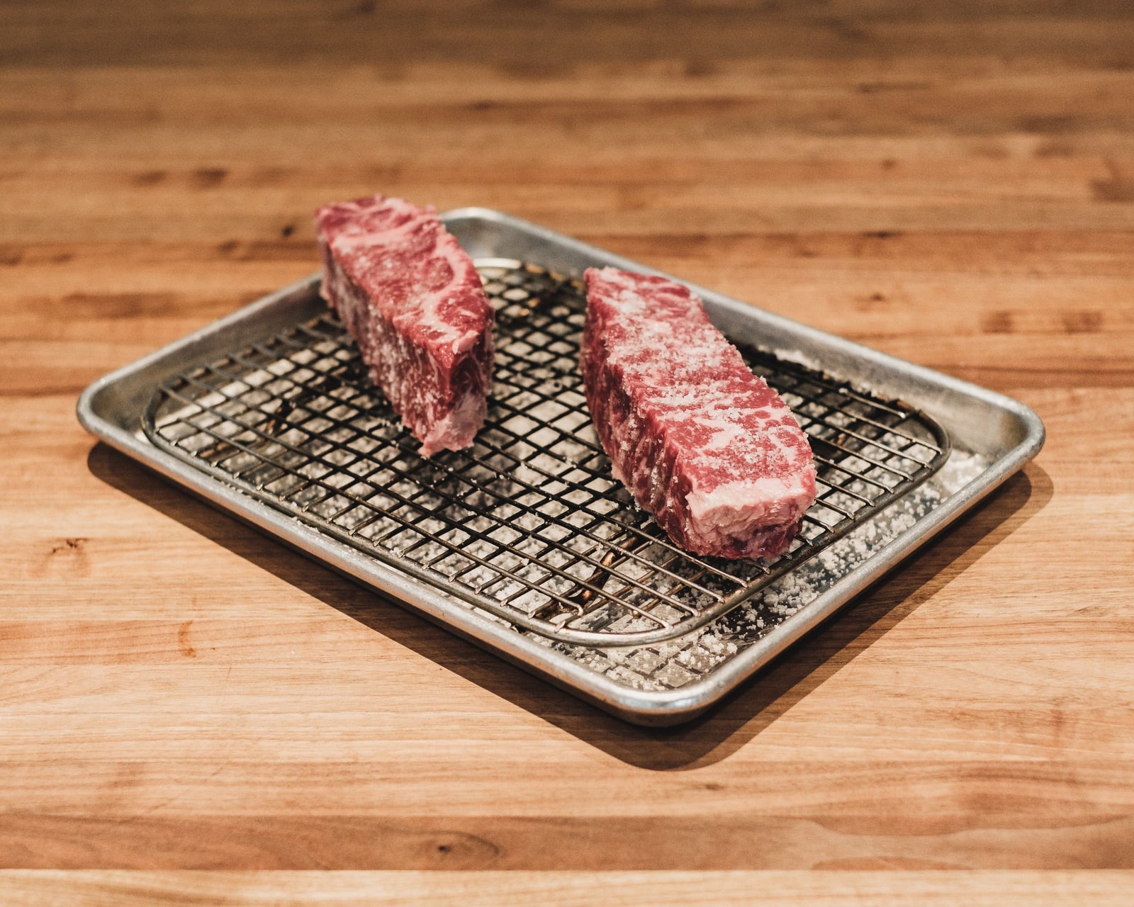 Two thick raw marbled steaks on a wire rack inside a small metal tray on a light wooden counter.