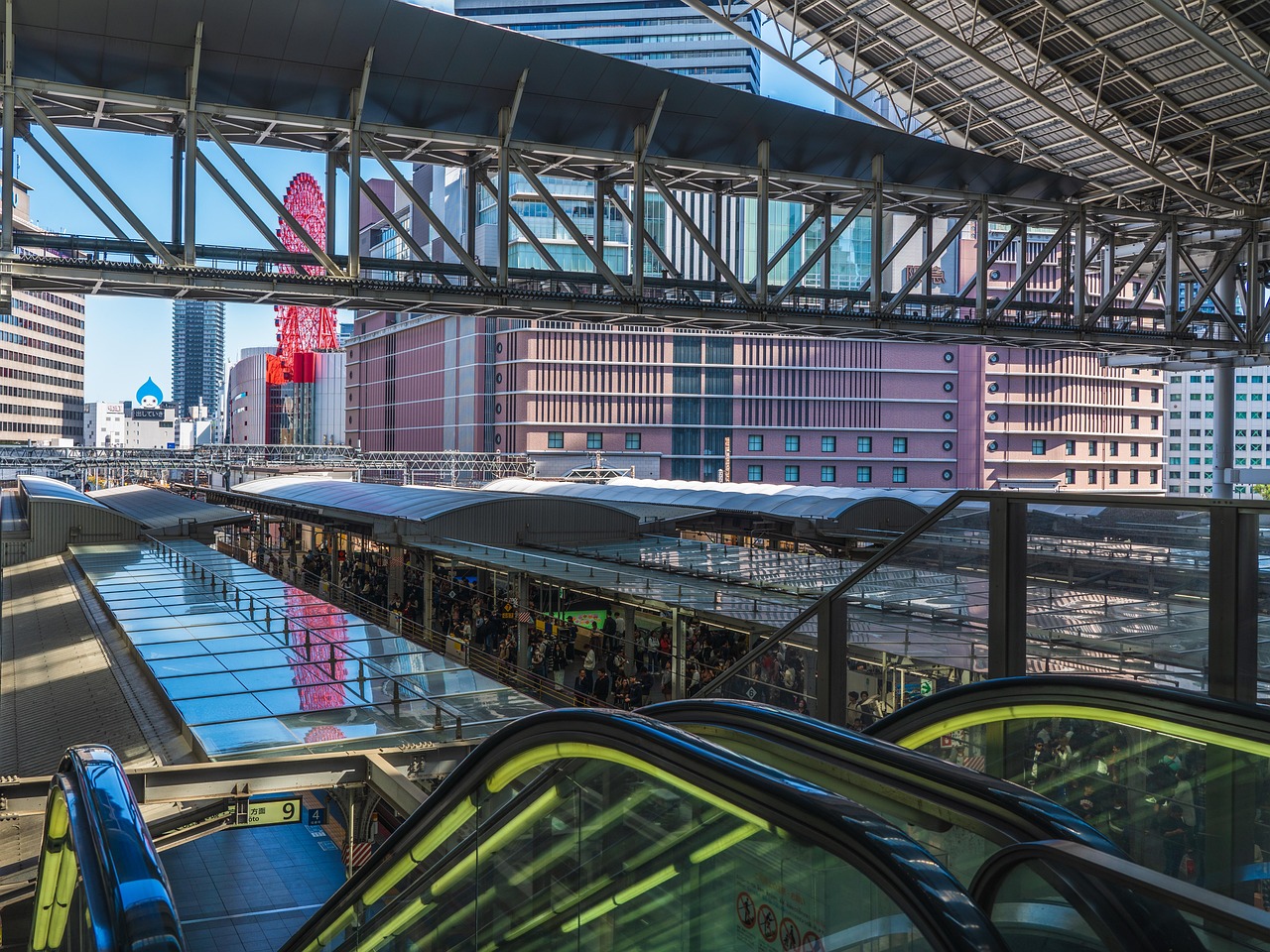 A large steel-and-glass roof over station platforms seen from an upper deck, escalators in front, office towers and a red Ferris wheel on a rooftop behind.