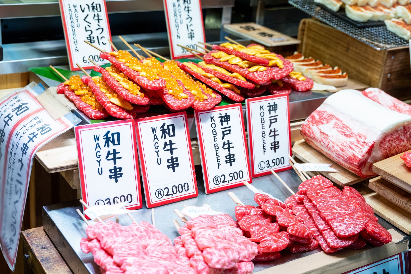A market counter of raw beef skewers and a marbled block, with handwritten cards labeling wagyu skewers and Kobe beef skewers in Japanese and English.