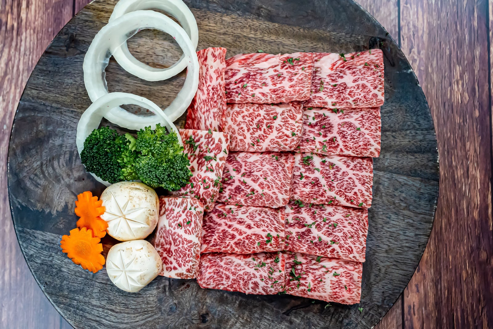 Thick raw beef slices with dense white marbling laid in rows on a round dark wooden platter, onion rings, broccoli and white mushrooms at the side.