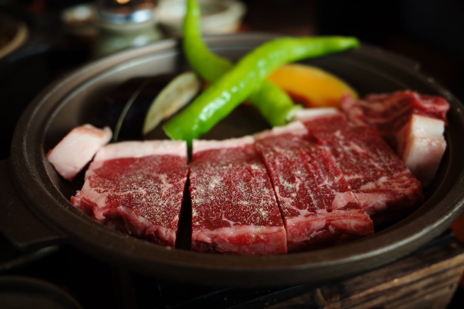 Cut slices of marbled beef in a dark iron dish, green shishito peppers and a slice of orange pepper on top.