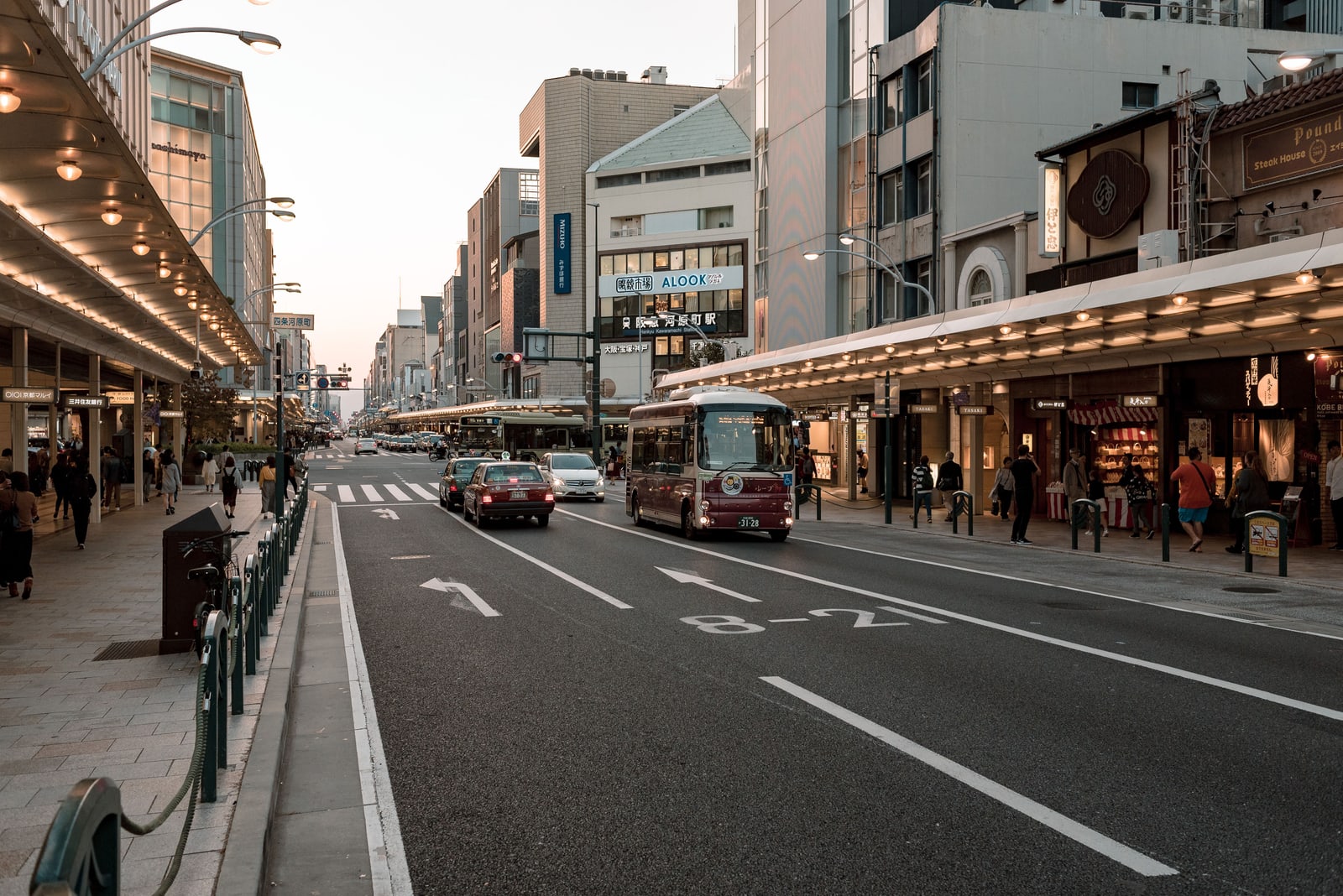 A wide downtown Kyoto avenue at dusk, a red-and-cream bus in the same lanes as the cars, covered sidewalks on both sides.