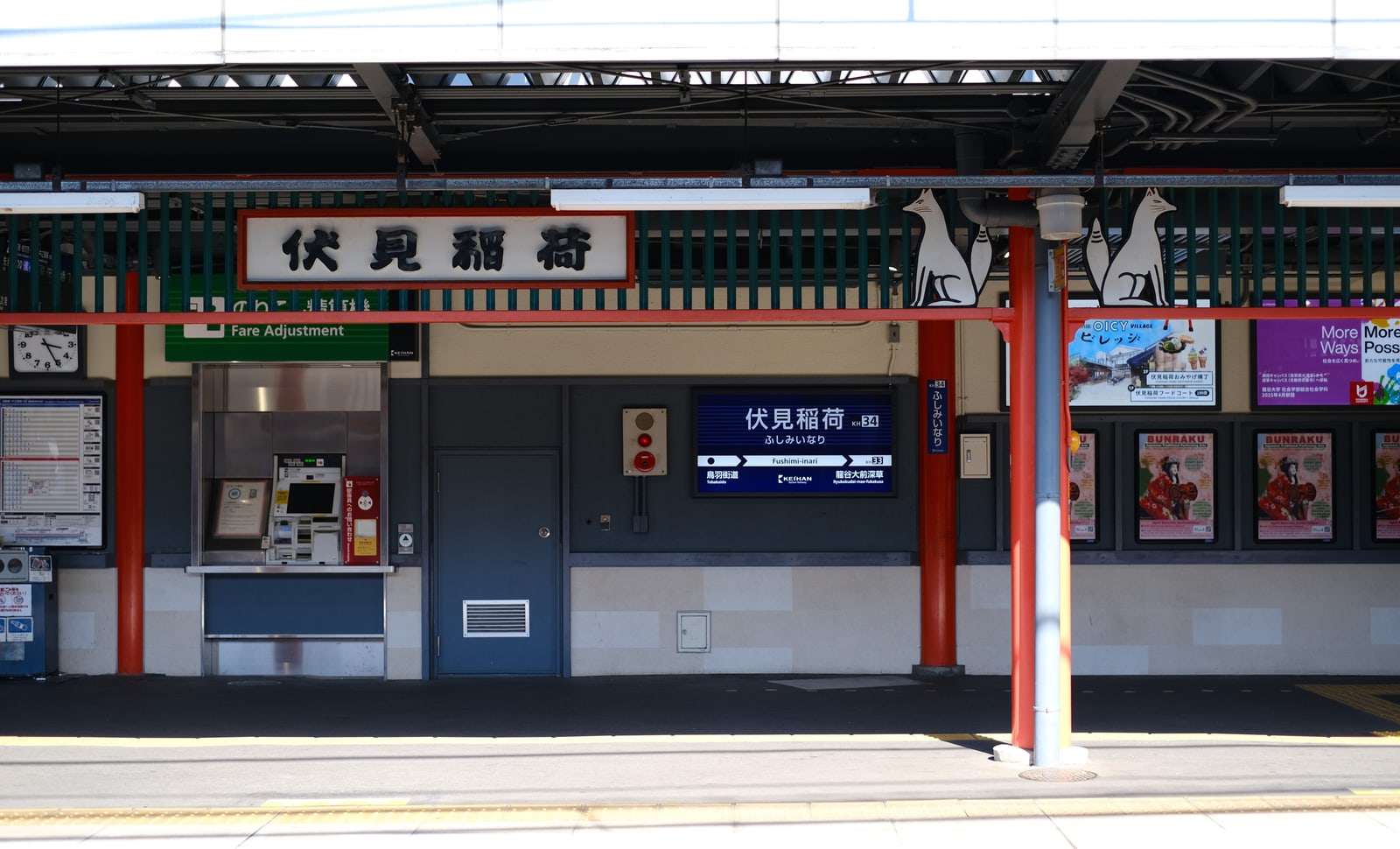 An empty open-air platform under a vermilion-pillared canopy, the station board reading Fushimi-Inari in Japanese, two white fox figures above it.
