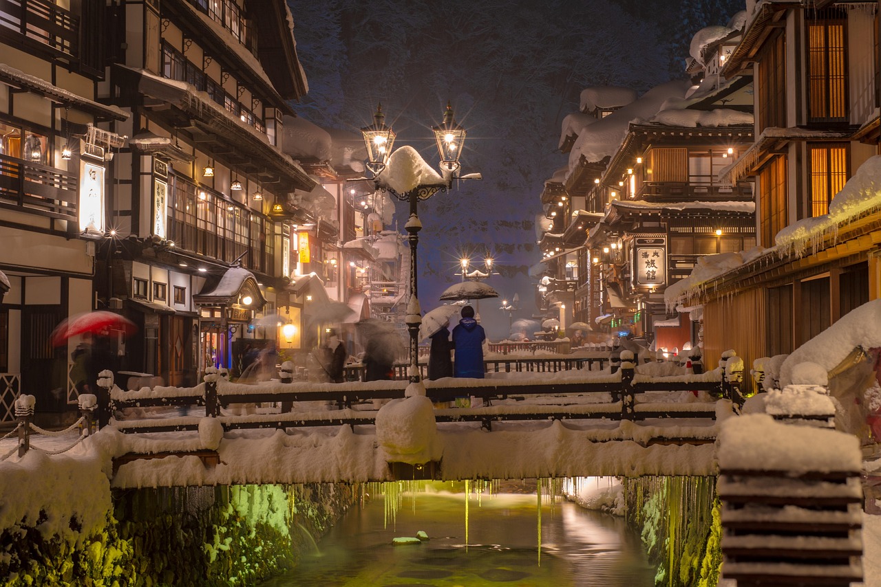 An onsen town street at night in heavy snow, wooden inns lit along a narrow stream and small figures with umbrellas crossing a bridge.