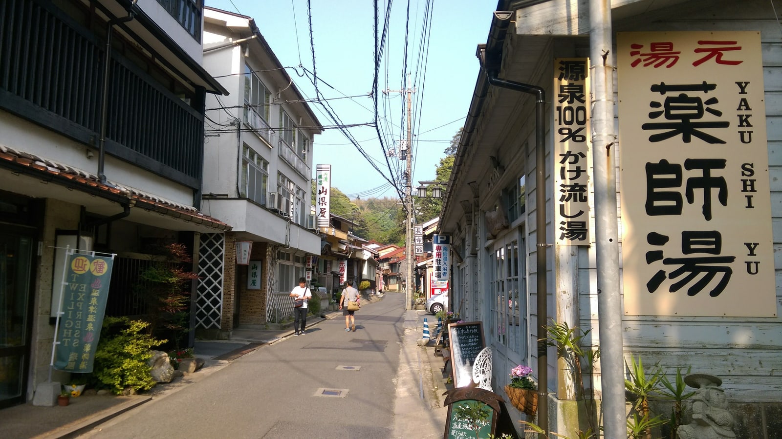 A quiet street of old two-story buildings, with a large wooden sign for a public bathhouse called Yakushi-yu on the right and two people walking in the distance.