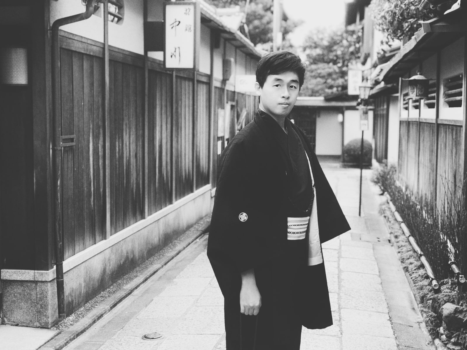 Black-and-white photo of a young man in a dark yukata standing in a stone-paved lane between wooden walls.