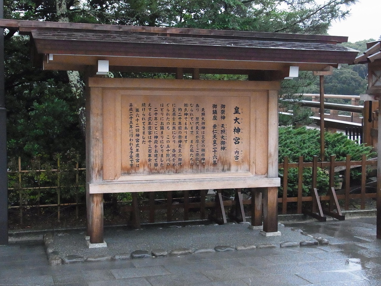 A roofed wooden notice board naming the Inner Shrine of Ise Jingu, with wet stone paving and a wooden bridge railing behind.
