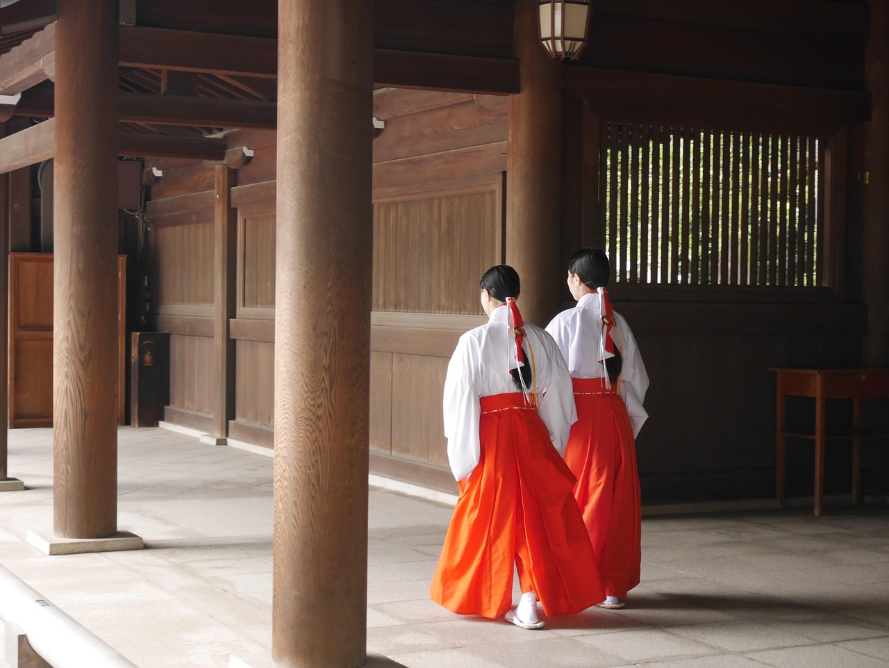 Two shrine attendants in white and red, seen from behind, walking along a wooden corridor between thick pillars.