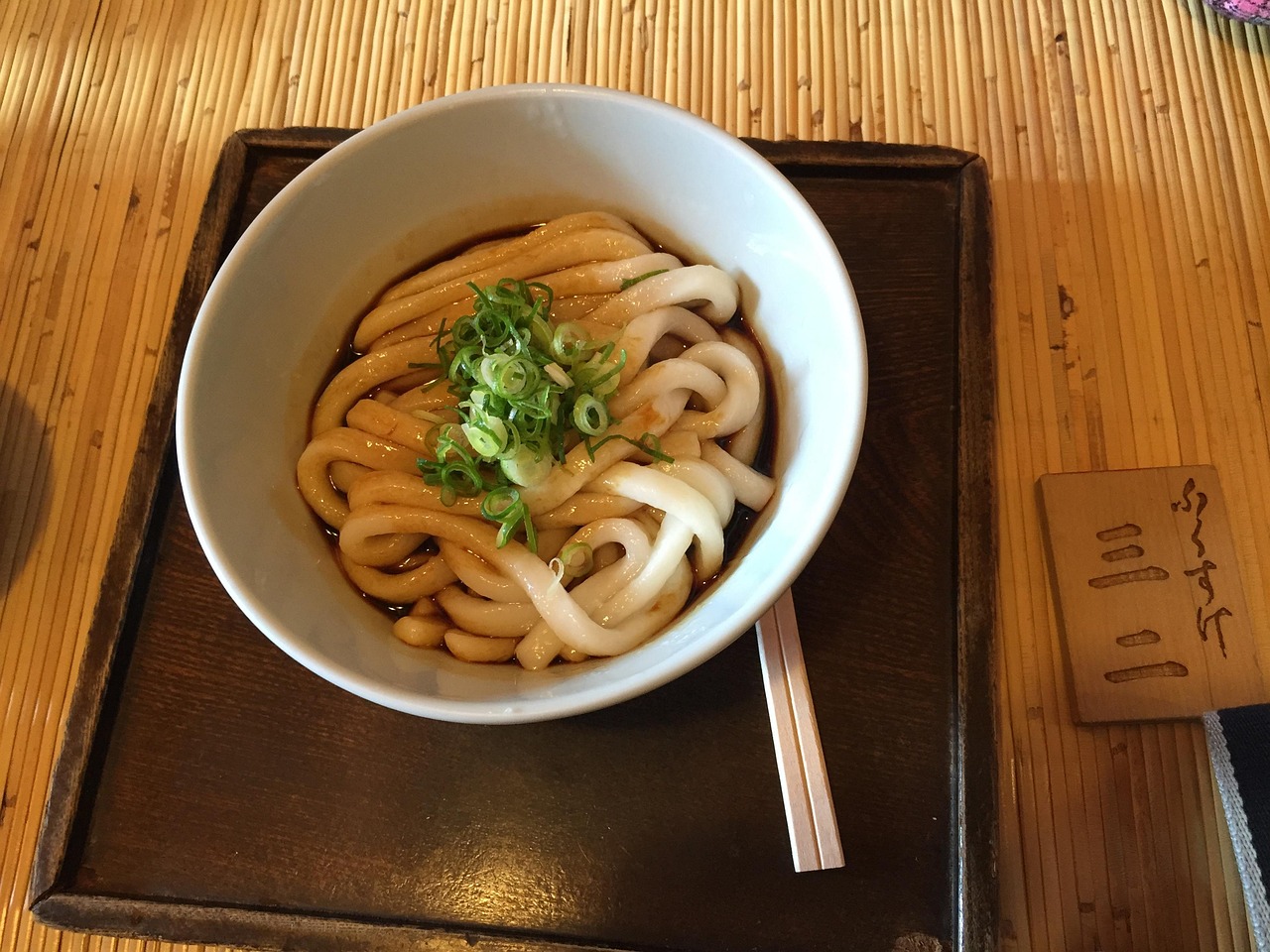 A bowl of thick udon in dark sauce with chopped green onion on a wooden tray, chopsticks beside it.