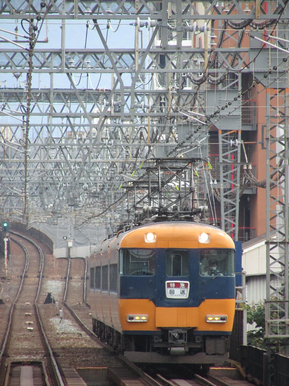 An older orange and navy Kintetsu limited express approaching under overhead wiring beside a tall building.