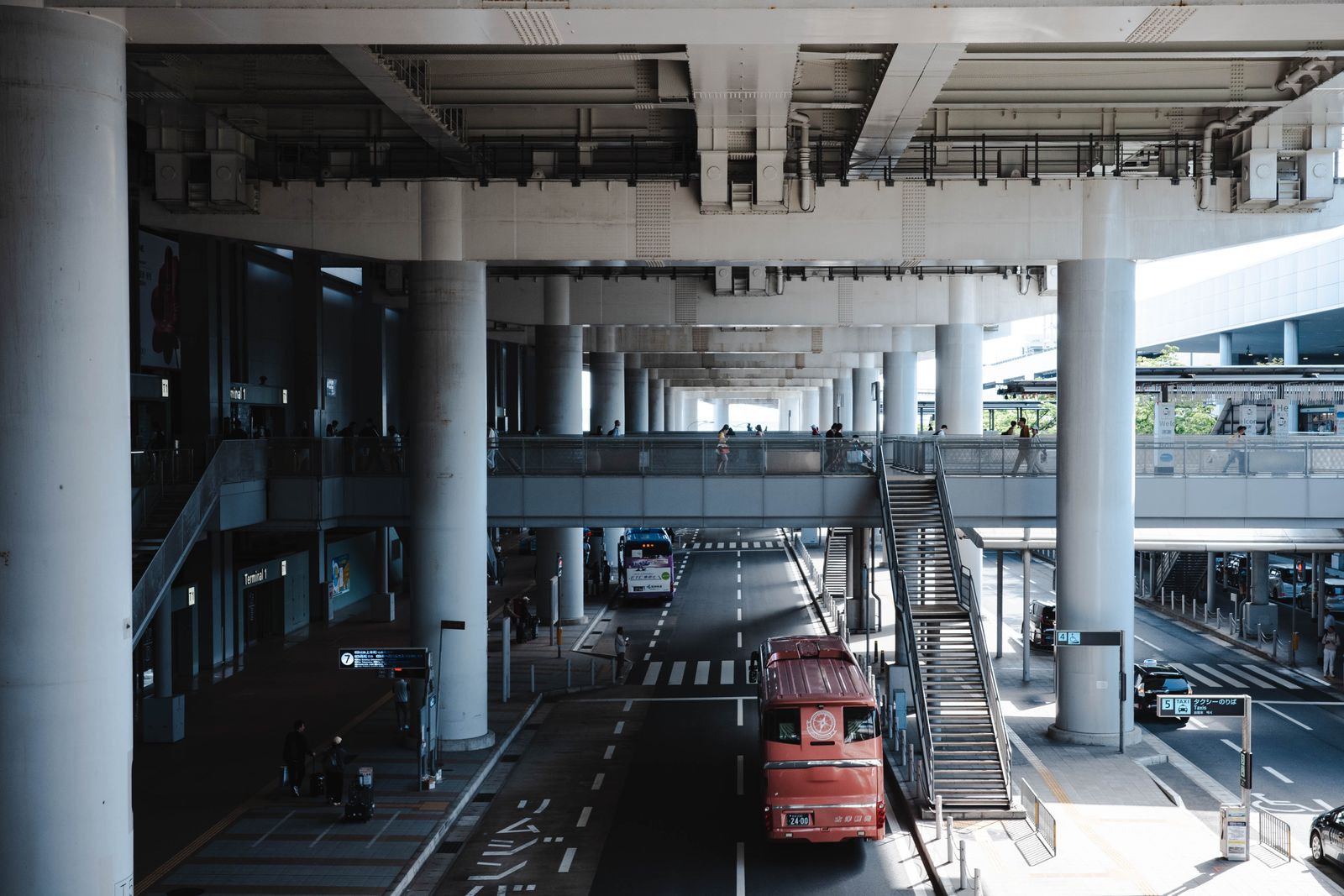 A covered road and footbridge under a concrete canopy at Kansai International Airport, a red bus below.