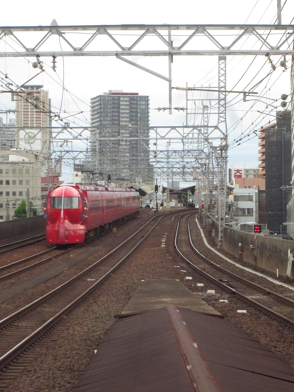 A red train with a rounded nose on a multi-track line between Osaka apartment blocks.