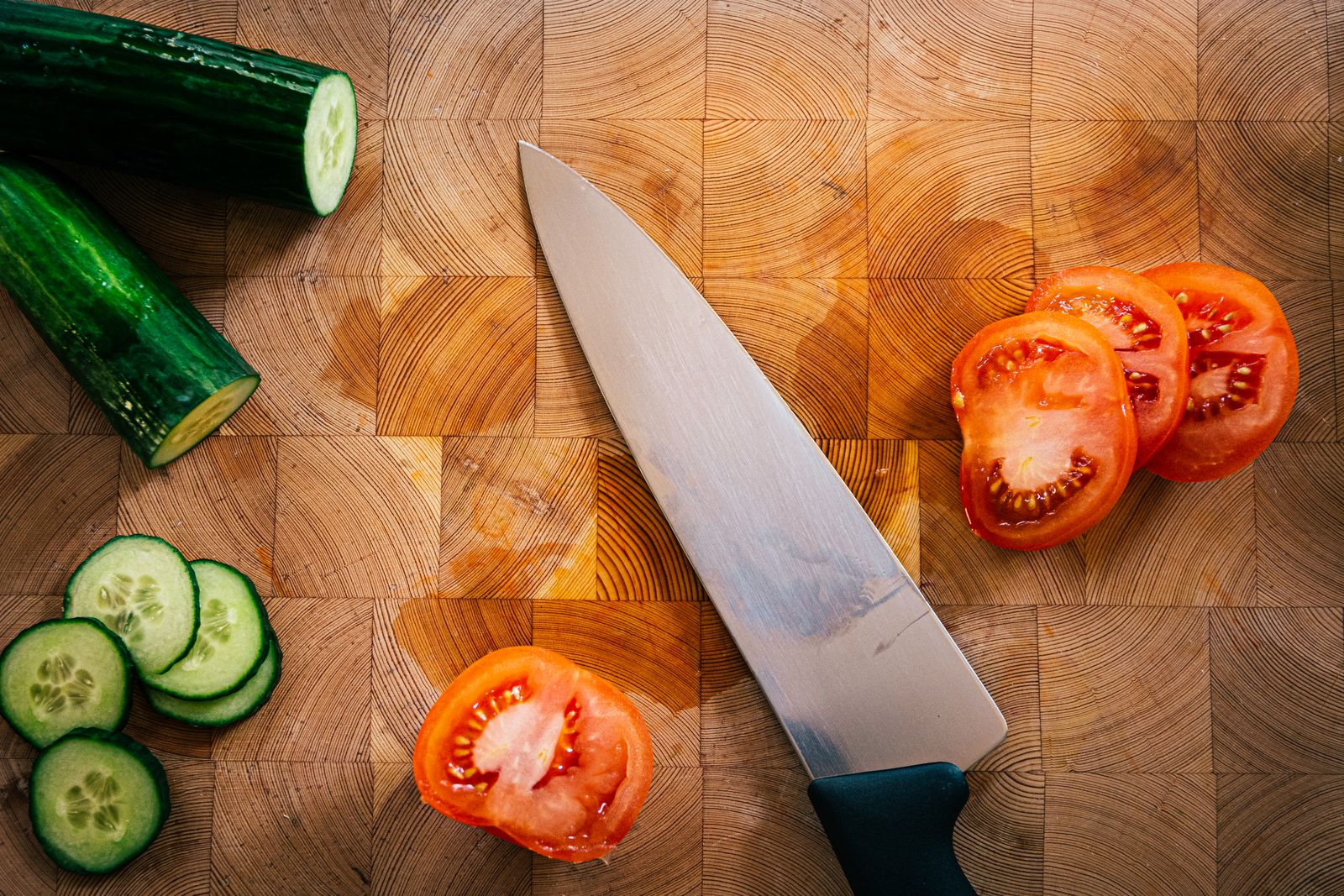 One kitchen knife on an end-grain wooden board, seen from above, with a cucumber and a tomato part-sliced beside it.