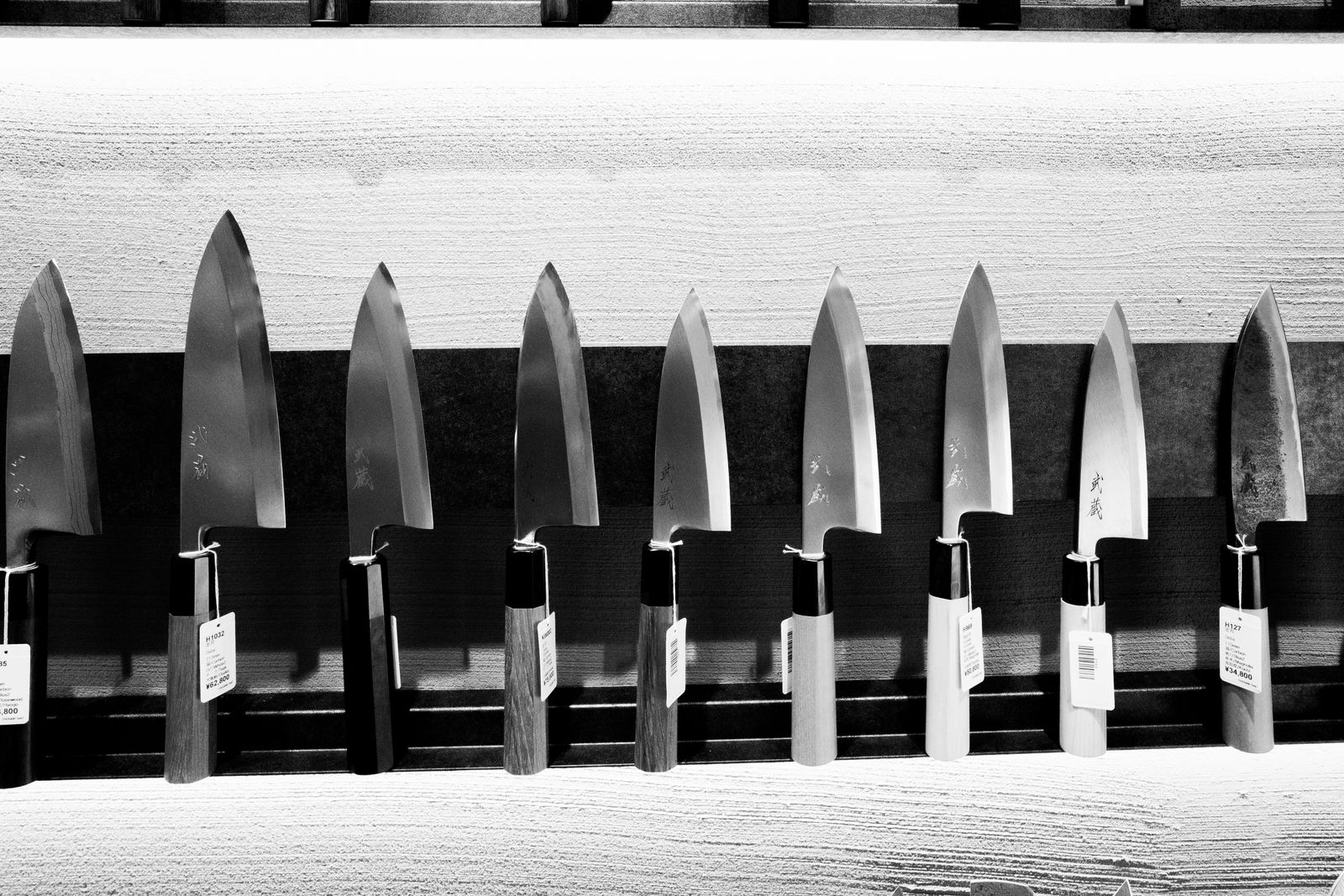 A row of Japanese knives hanging on a shop rack, blades engraved with kanji and tags showing yen prices.