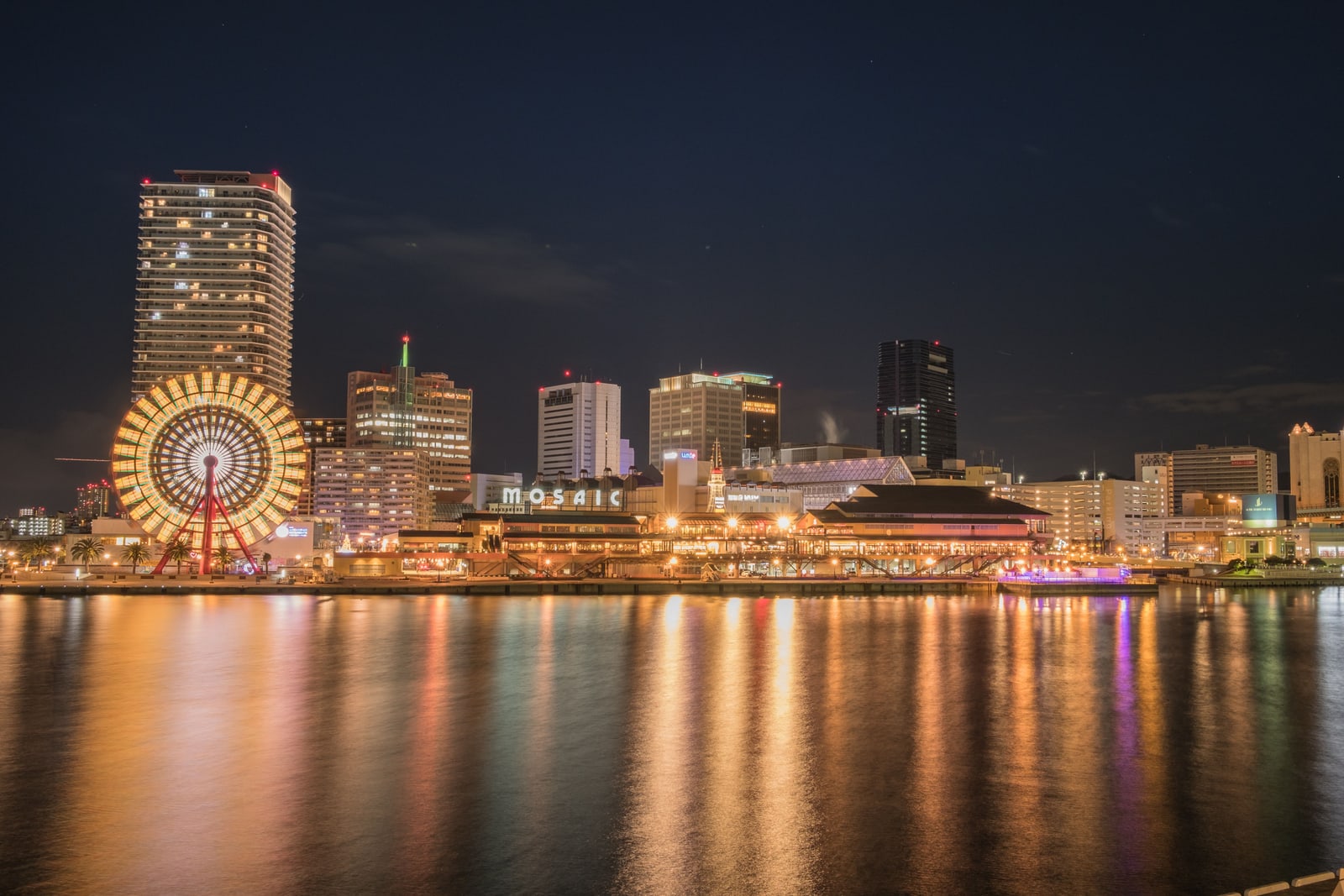 At night across still water: a lit waterfront complex with a MOSAIC sign, a large lit Ferris wheel beside a tall apartment tower, amber lights reflected in the water