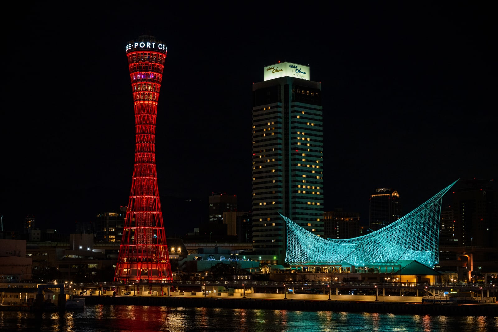At night across harbour water: the red lattice Port Tower lit red, a tall hotel tower with a lit rooftop sign, and the white sail-shaped steel-frame roof of the Maritime Museum lit blue-green