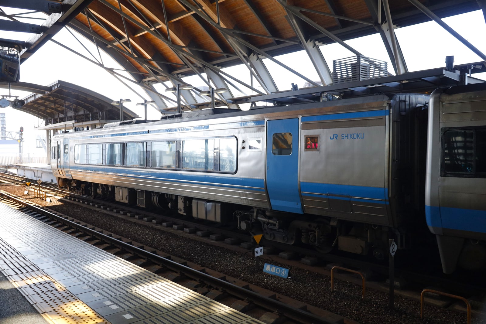 A silver and blue JR Shikoku train beside a platform under a high curved wooden-and-steel roof