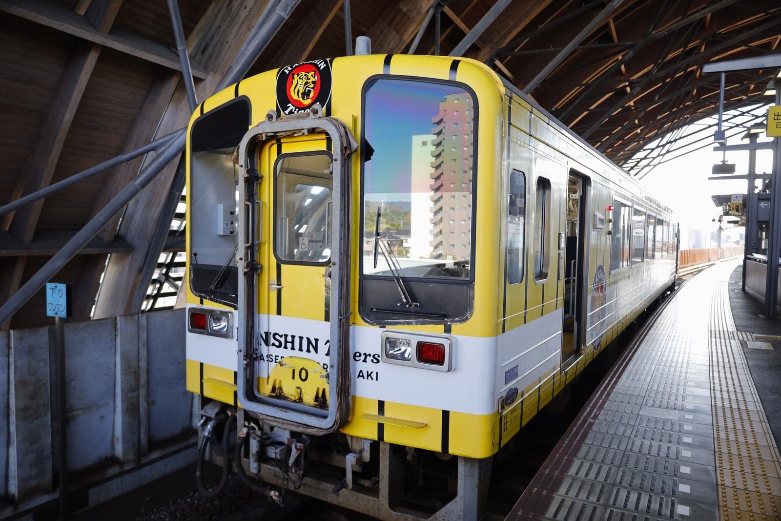 The front of a yellow, white and black striped railcar with a tiger-head logo, stopped beside a platform under a curved wooden-and-steel roof