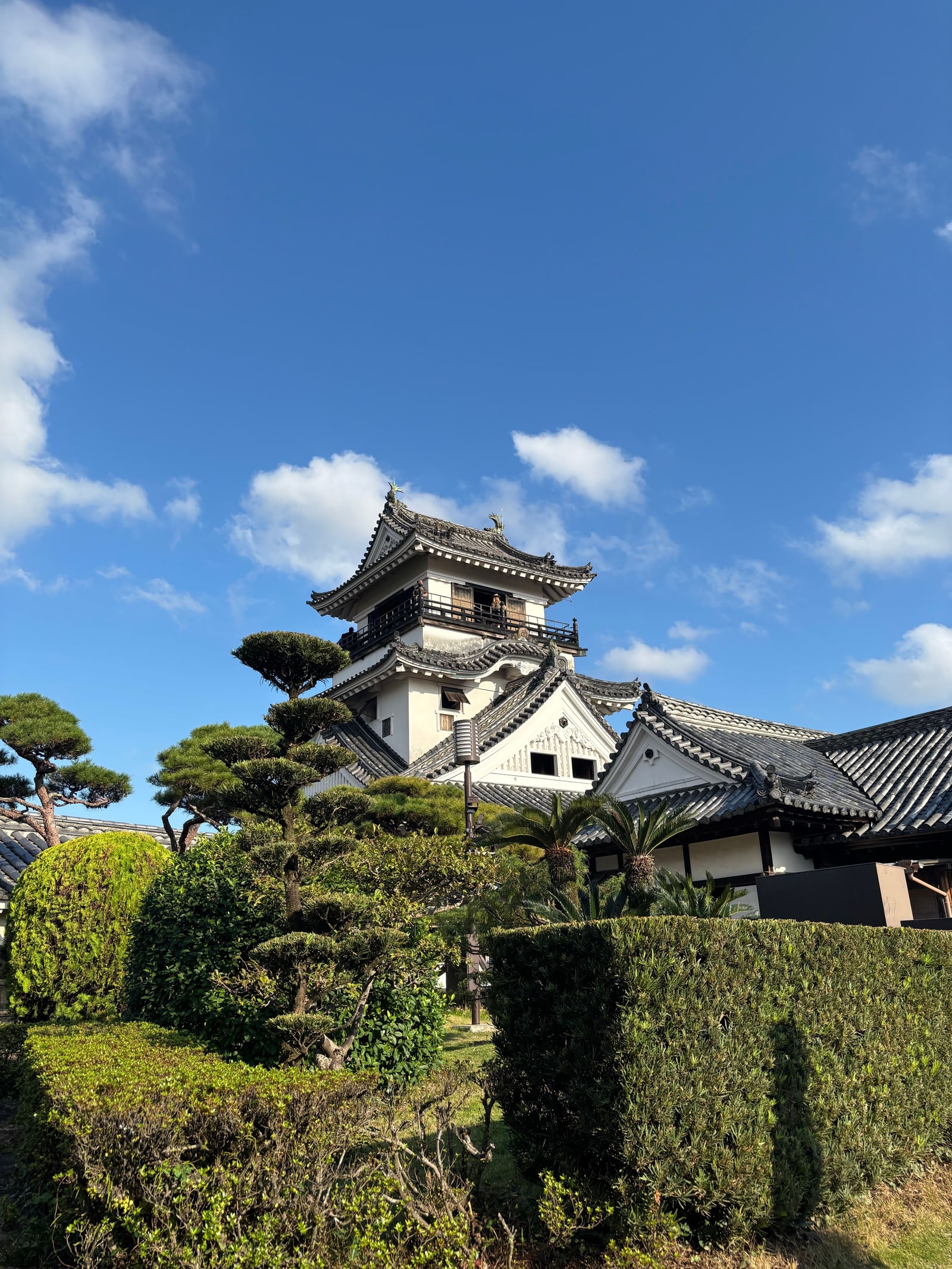 A white castle keep with a wooden balcony behind clipped pines and hedges, strong blue sky with white clouds