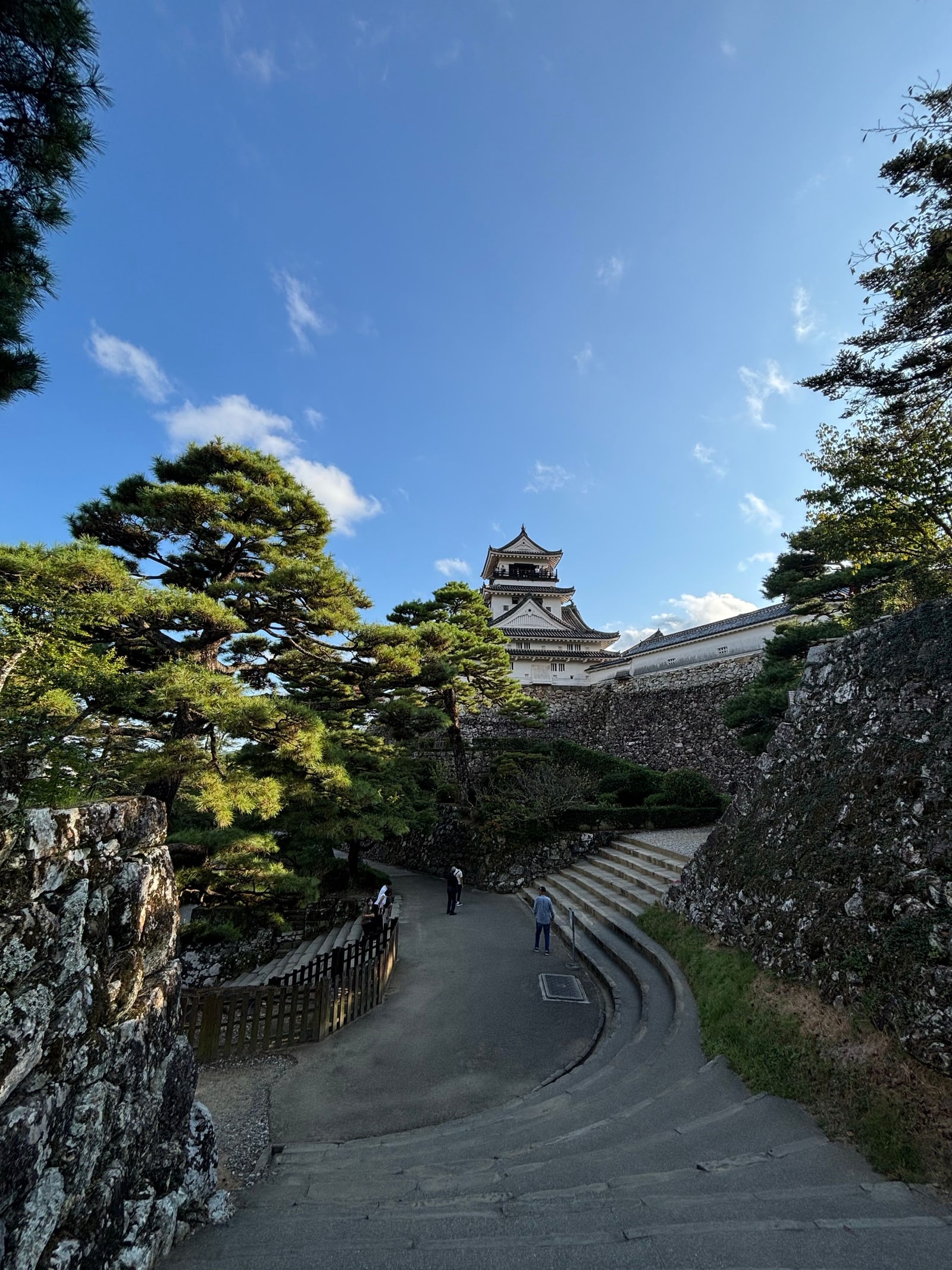 A white castle keep at the top of a curving stone-paved path between stone walls and pines, three visitors on the path, deep blue sky