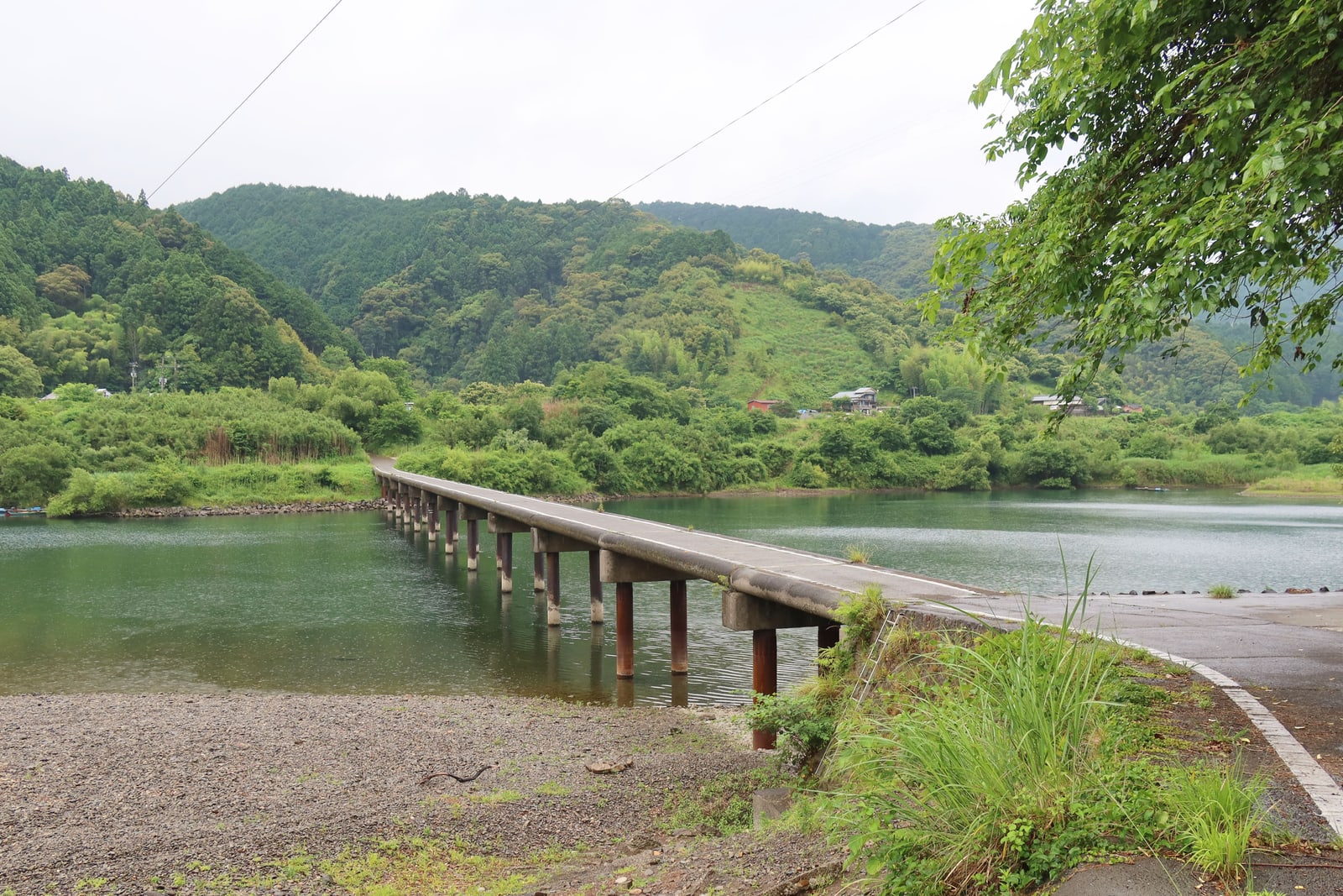 A long low concrete bridge with no railings crossing a green river, forested hills beyond, overcast sky