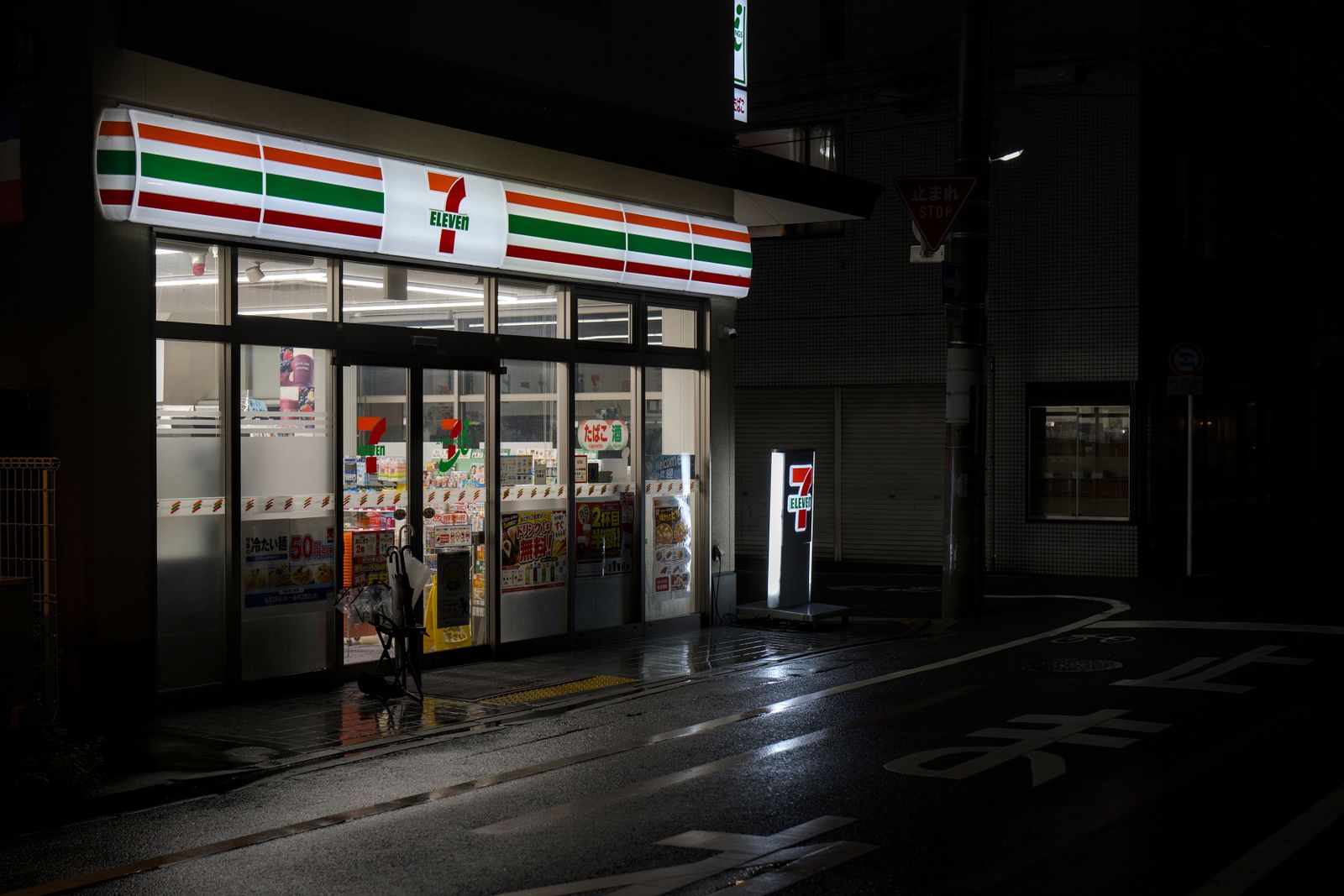 A 7-Eleven lit up on a wet, empty street in Kyoto at night, with a Japanese stop sign on the pole beside it.